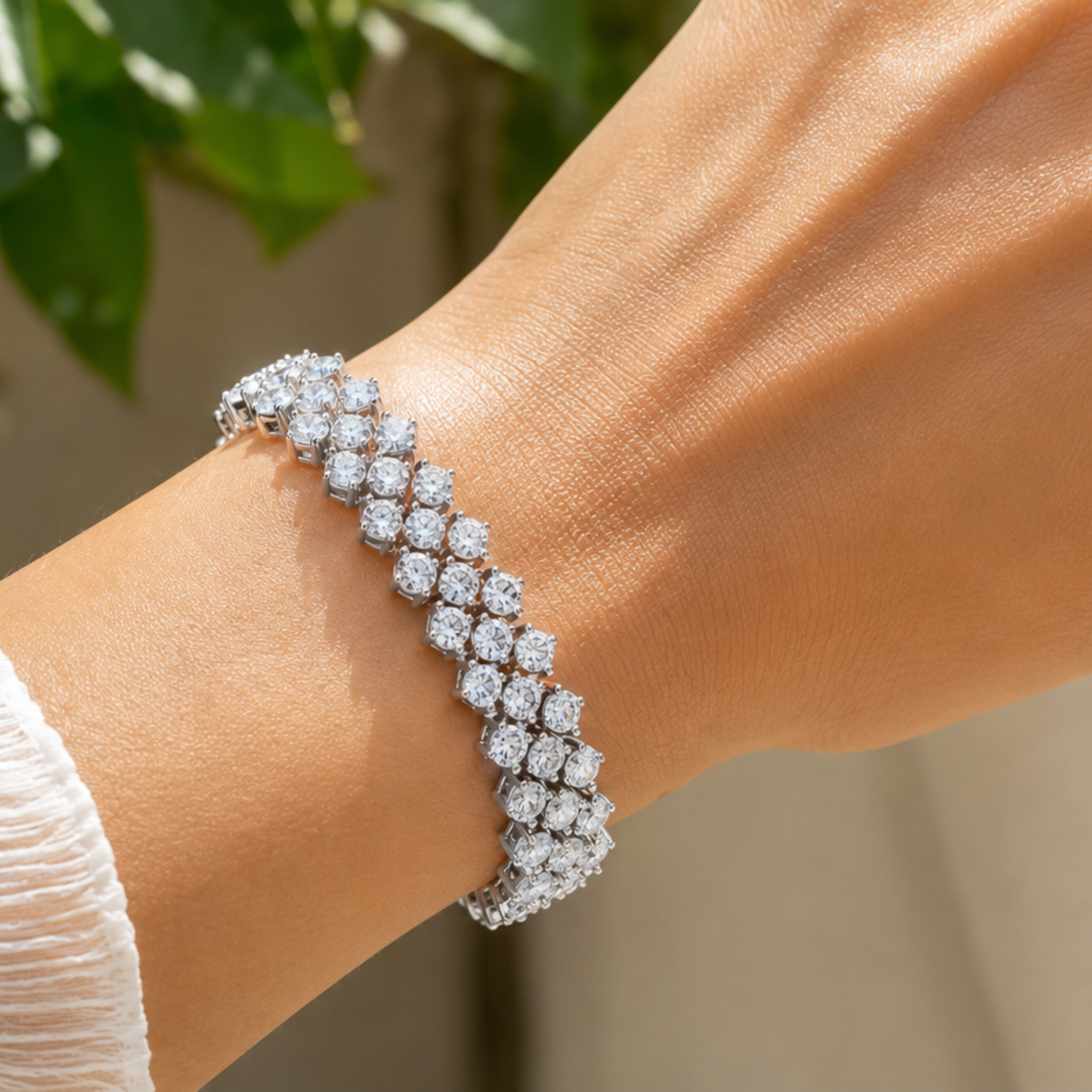 A close-up of a double-row diamond tennis bracelet on a woman's wrist, set in silver-toned metal with a soft-focus background of green leaves.