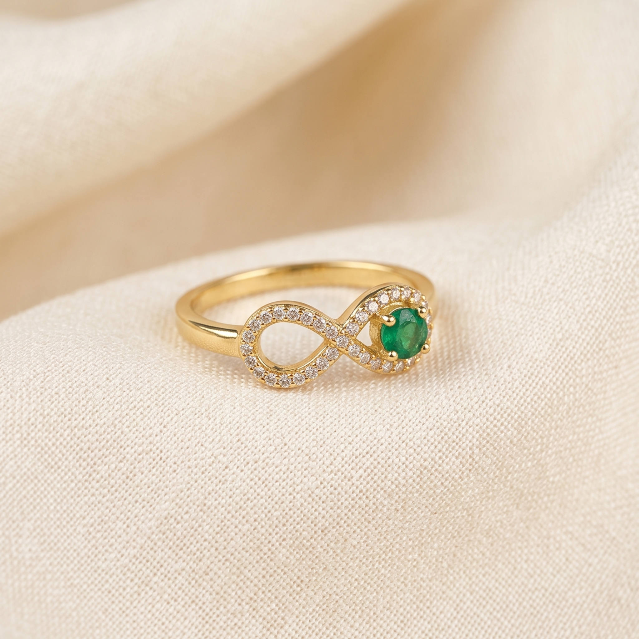 A gold infinity ring featuring a round green emerald center stone and a halo of small clear crystals, resting on a soft beige fabric background.