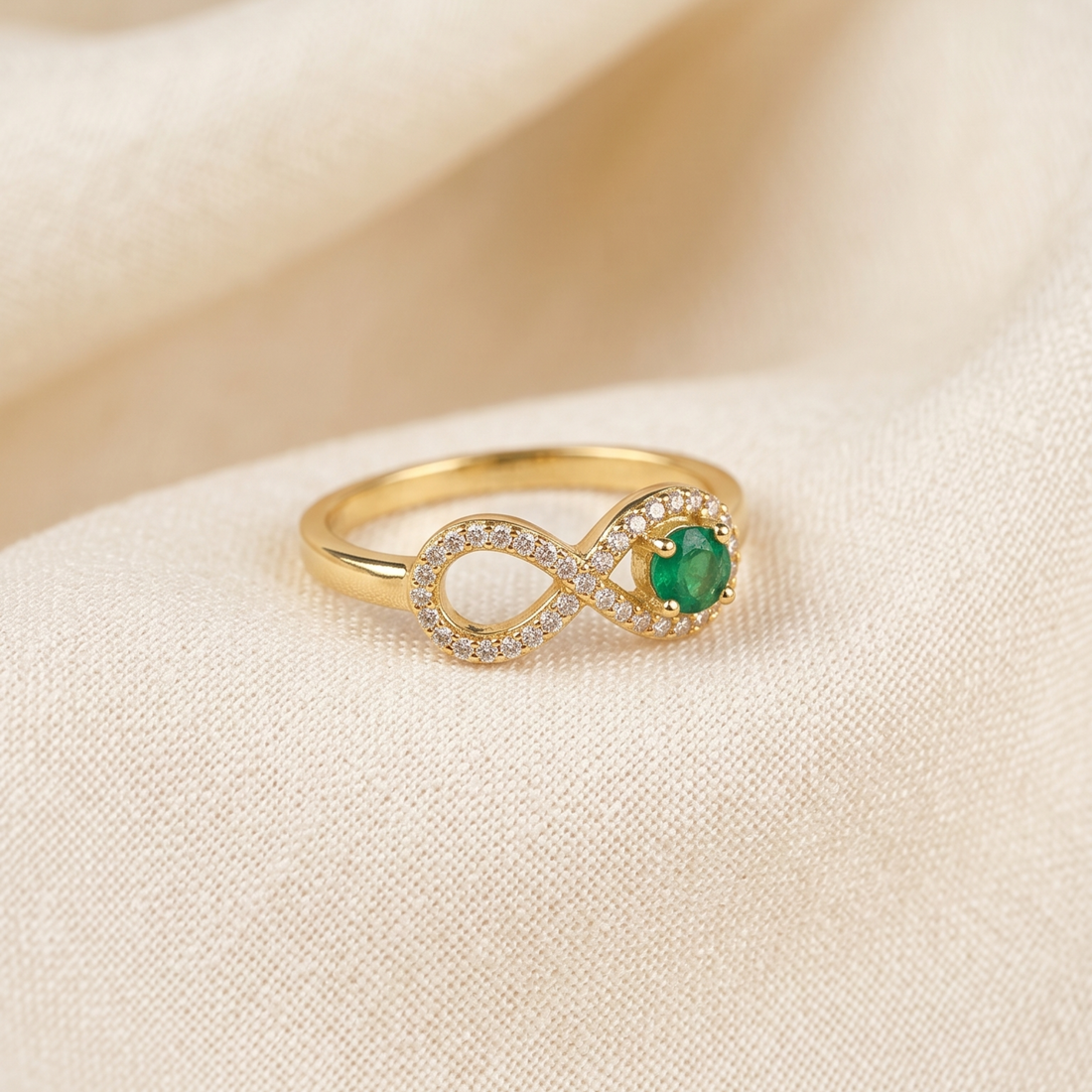 A gold infinity ring featuring a round green emerald center stone and a halo of small clear crystals, resting on a soft beige fabric background.