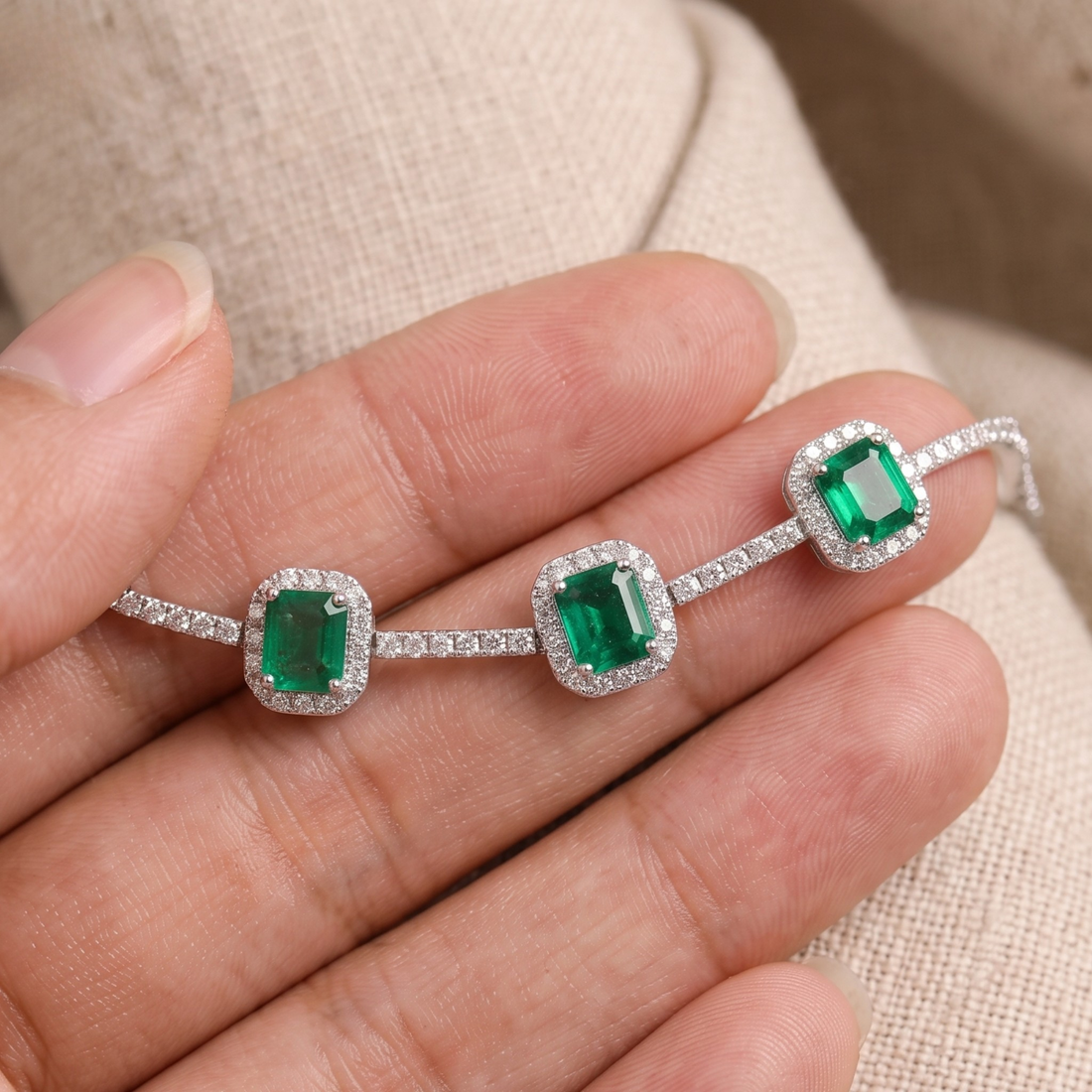 A close-up of a silver-toned bracelet with three emerald-cut green gemstones, each surrounded by a halo of small clear stones, resting on a person's fingers against a beige fabric background.