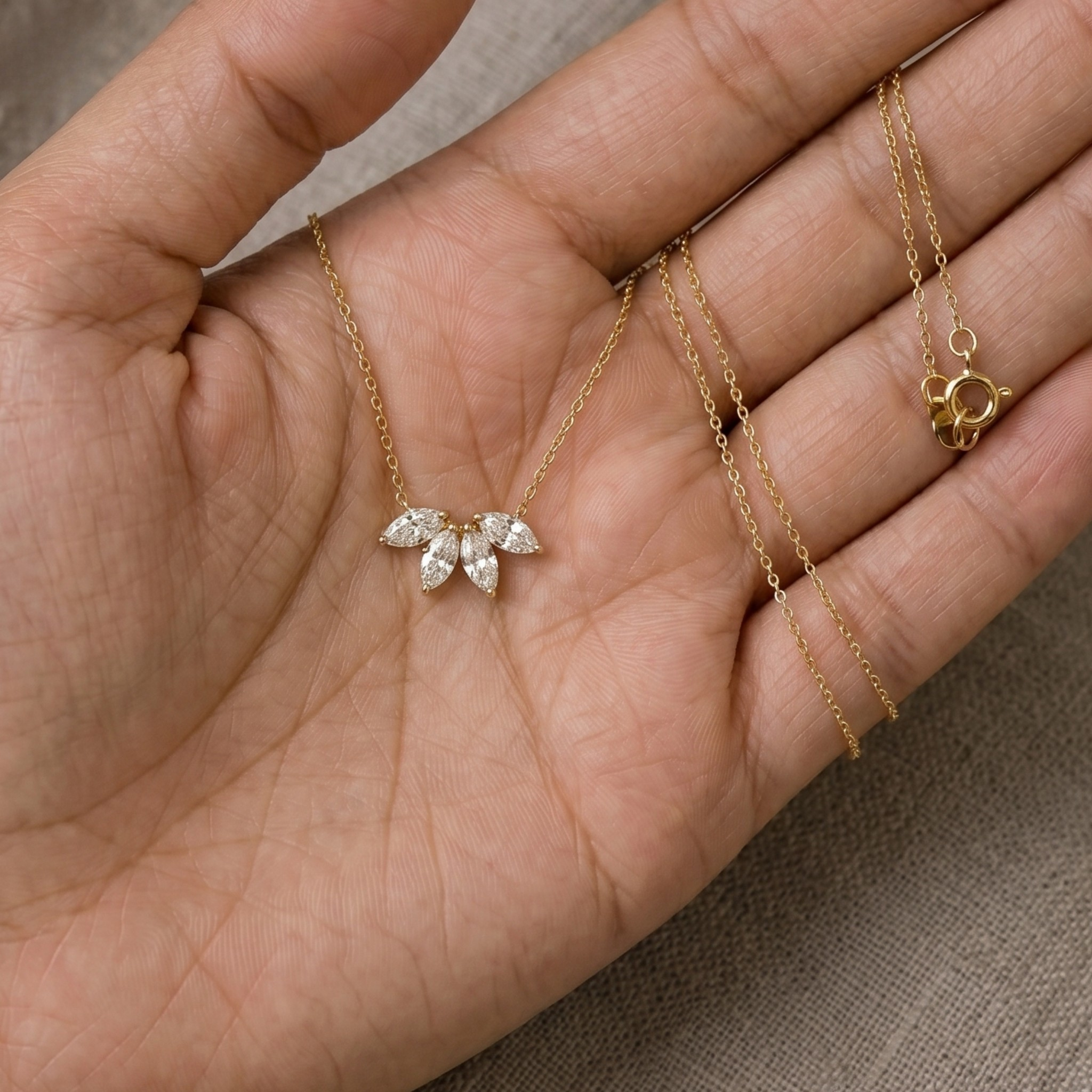 A delicate gold necklace with a small leaf cluster pendant made of clear marquise-cut stones, displayed on an open hand. The chain is draped across the fingers, and the clasp is shown separately on the right.