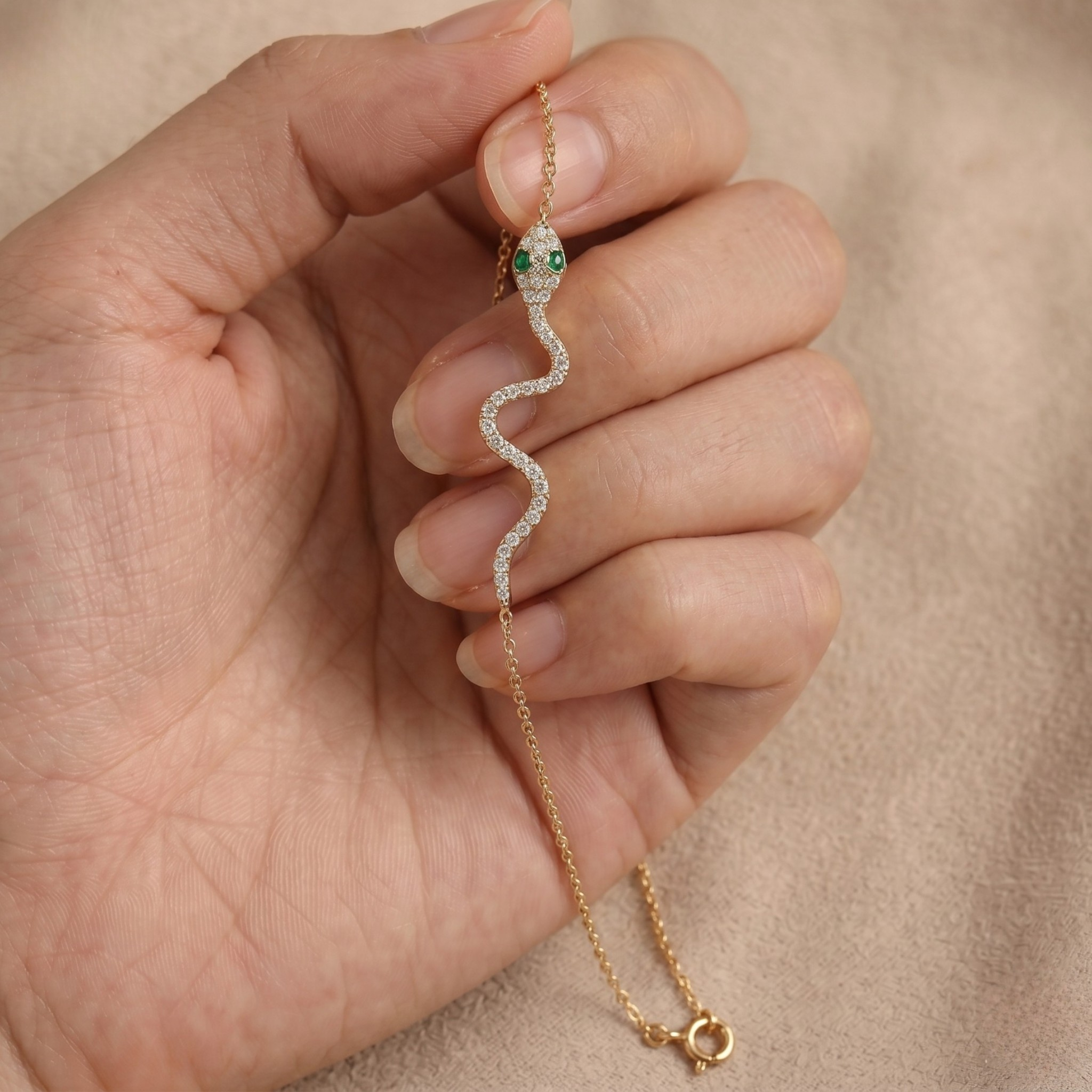 A gold snake bracelet with a green emerald-studded head and clear pavé body, held in a person's hand against a beige fabric background.