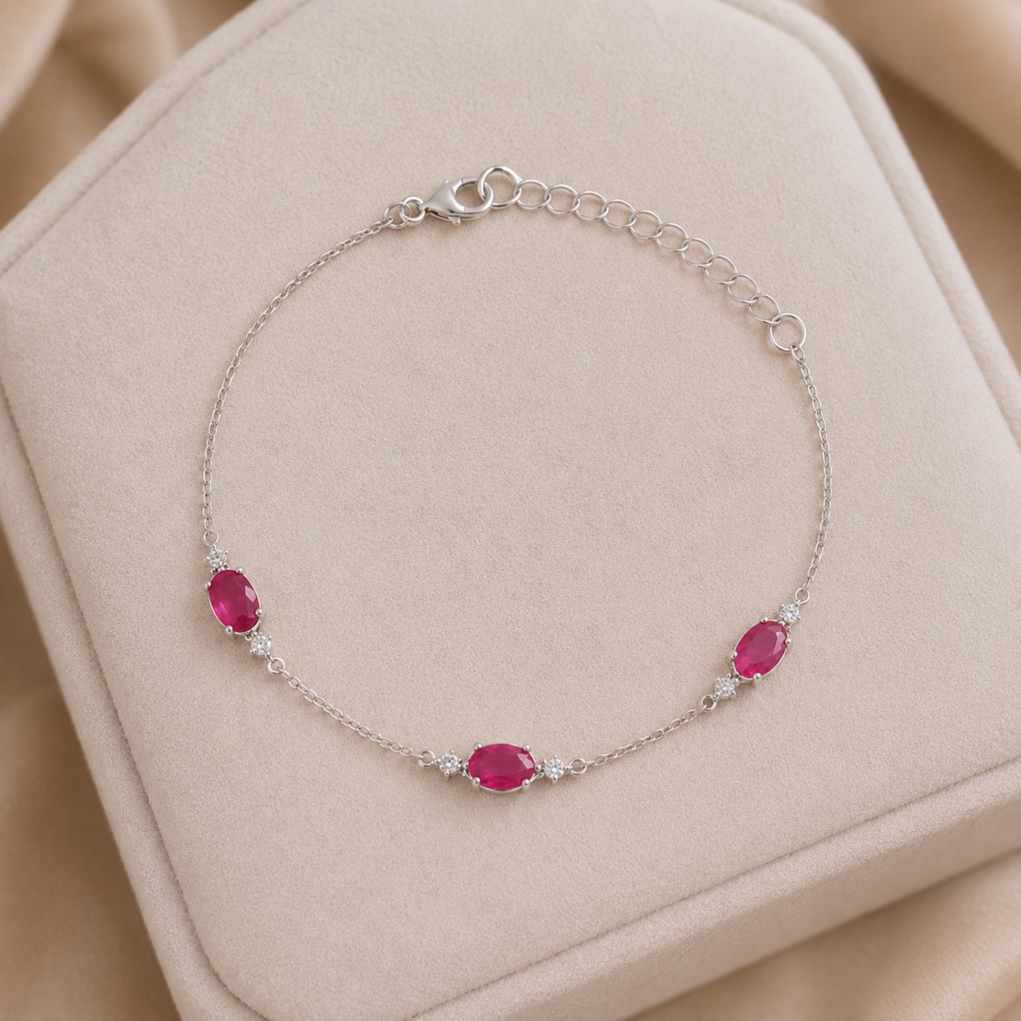 A delicate silver chain bracelet with three oval pink gemstones and small clear accent stones, displayed on a beige jewelry box.
