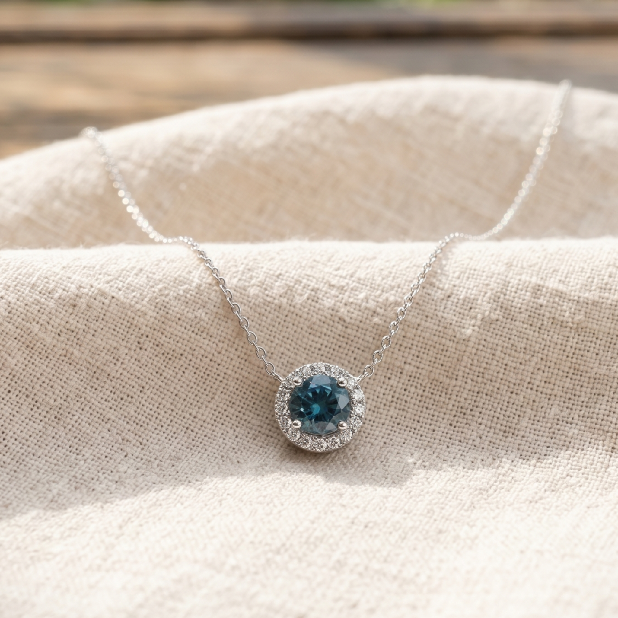A silver necklace with a round halo pendant featuring a deep blue gemstone, displayed on a textured beige linen fabric.