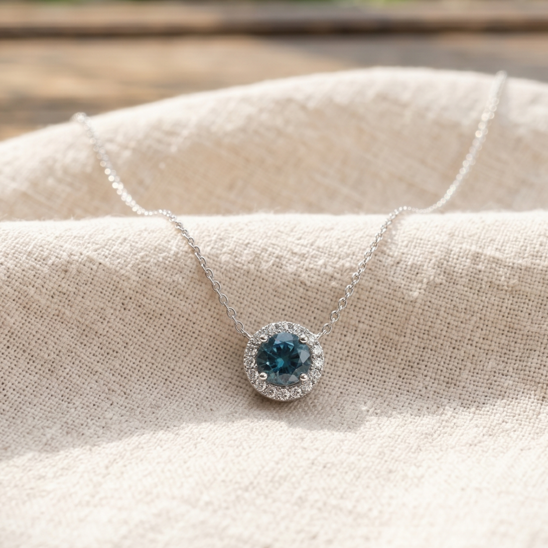 A silver necklace with a round halo pendant featuring a deep blue gemstone, displayed on a textured beige linen fabric.