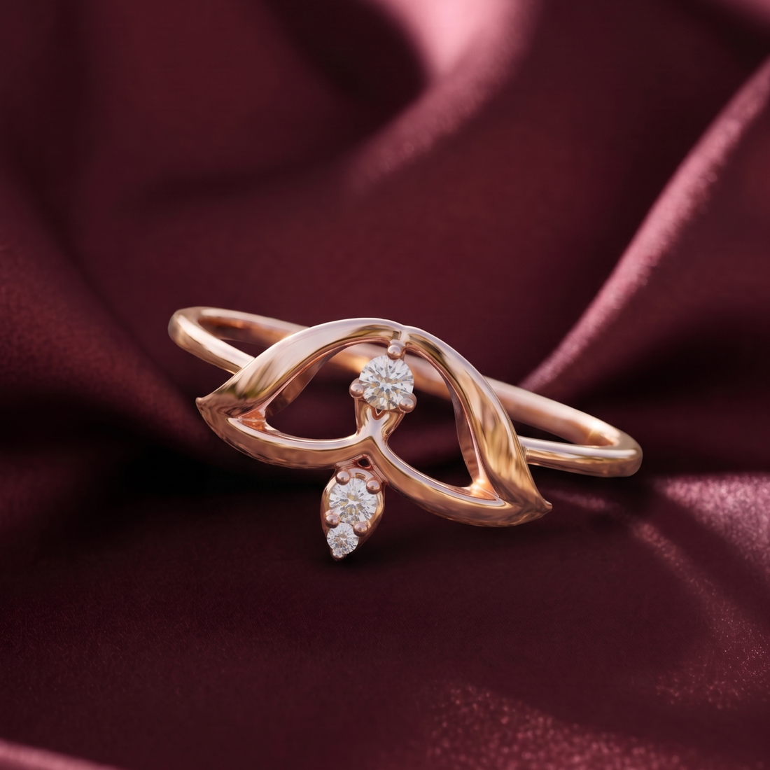 A rose gold ring with a sculpted, flowing openwork design and two clear gemstones, displayed on draped burgundy satin fabric.
