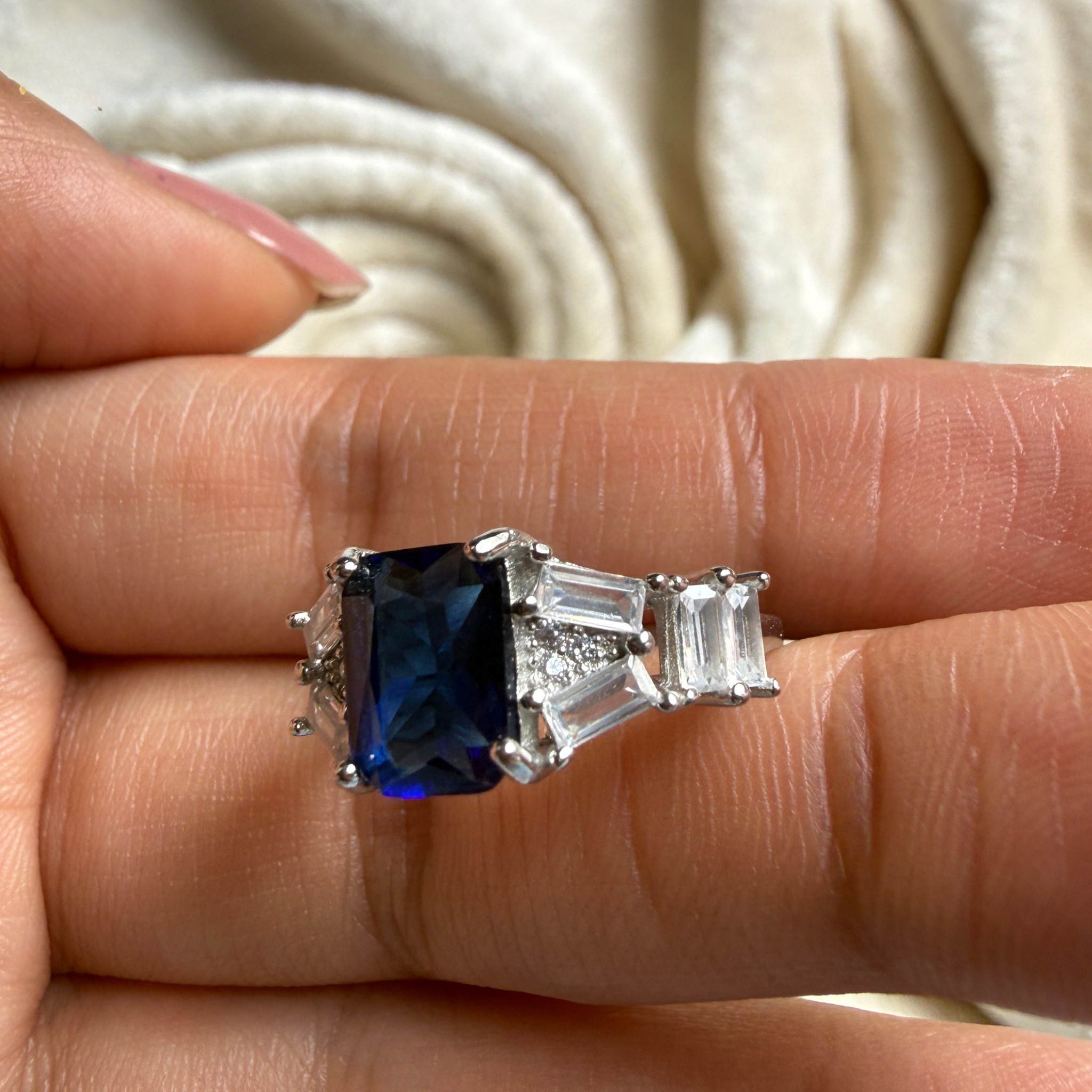 A silver ring featuring a rectangular dark blue sapphire center stone and two baguette-cut clear stones on each side, shown worn on a person's finger.