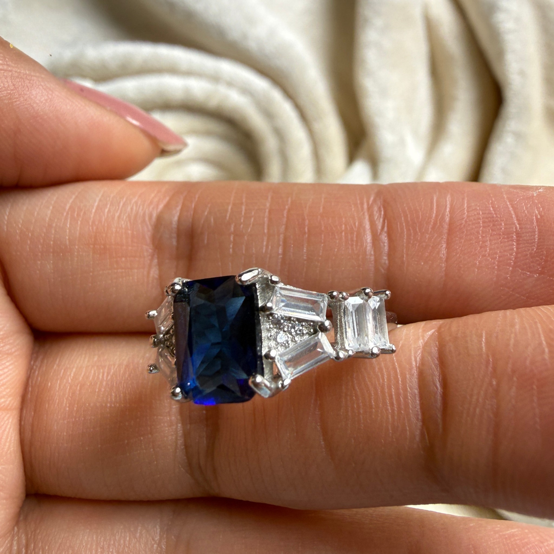 A silver ring featuring a rectangular dark blue sapphire center stone and two baguette-cut clear stones on each side, shown worn on a person's finger.