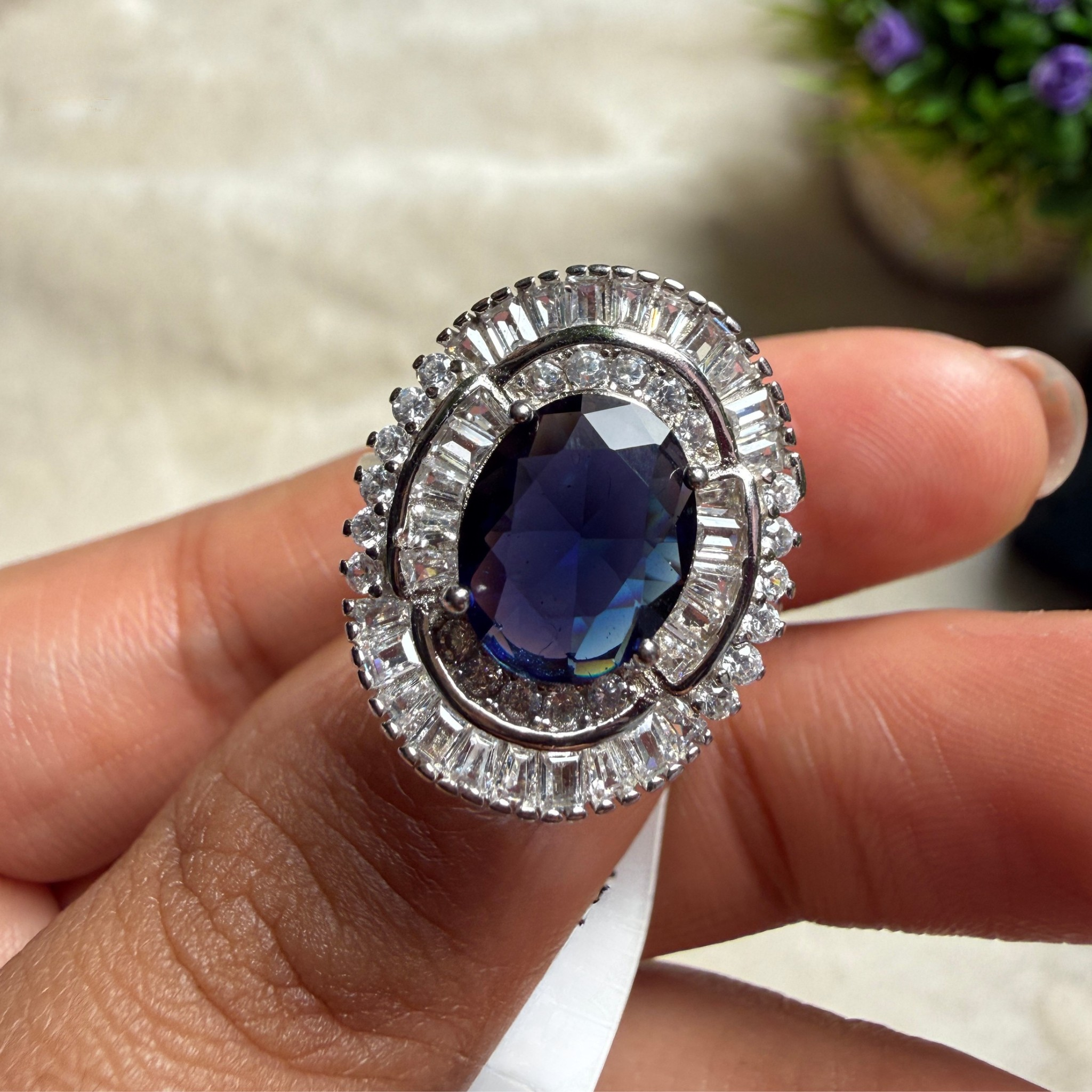 A close-up of a hand holding a silver-toned oval ring featuring a deep blue oval center stone surrounded by clear baguette and round-cut stones.