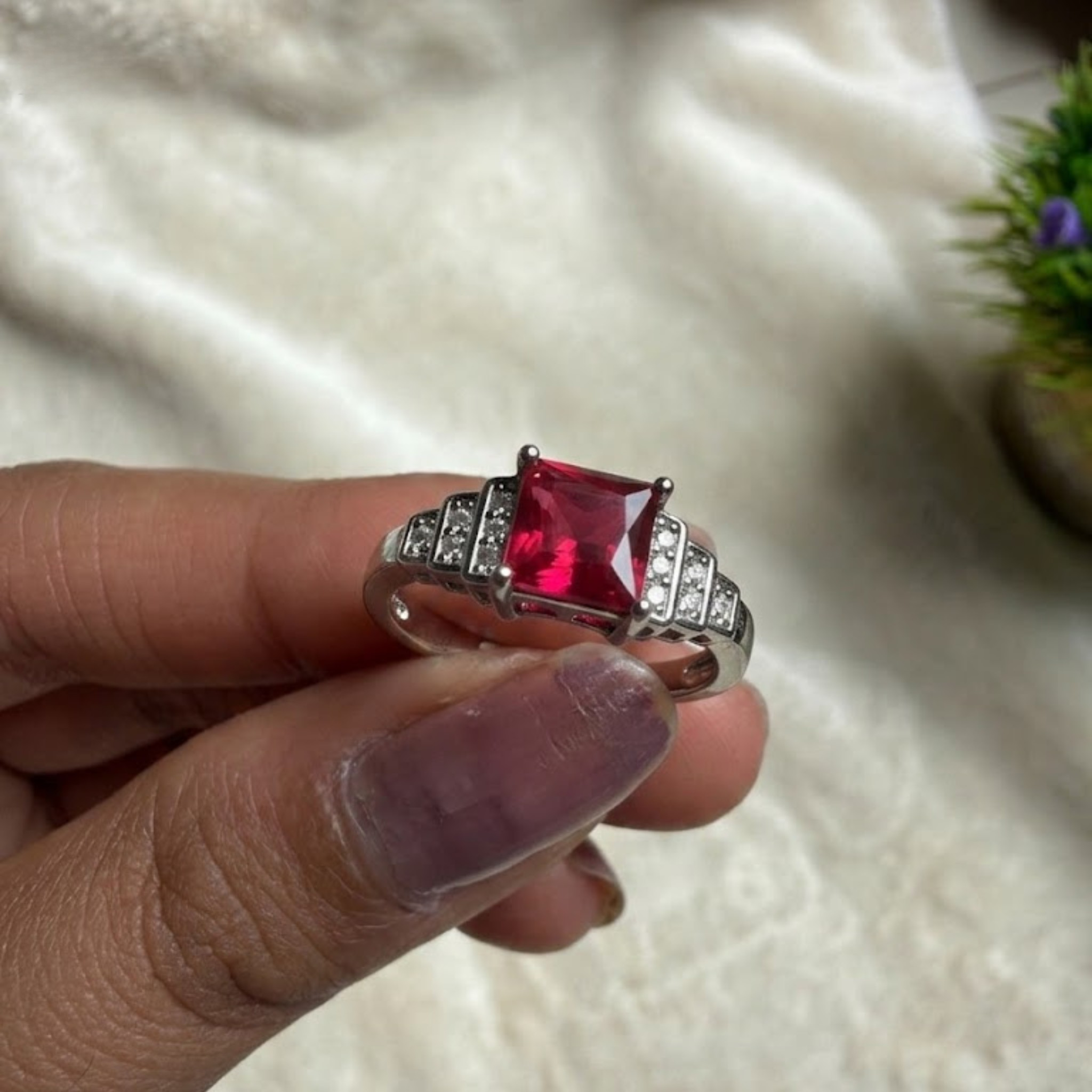 A silver ring with a square-cut red gemstone and small clear stones on the band, held between fingers against a soft white fabric background.