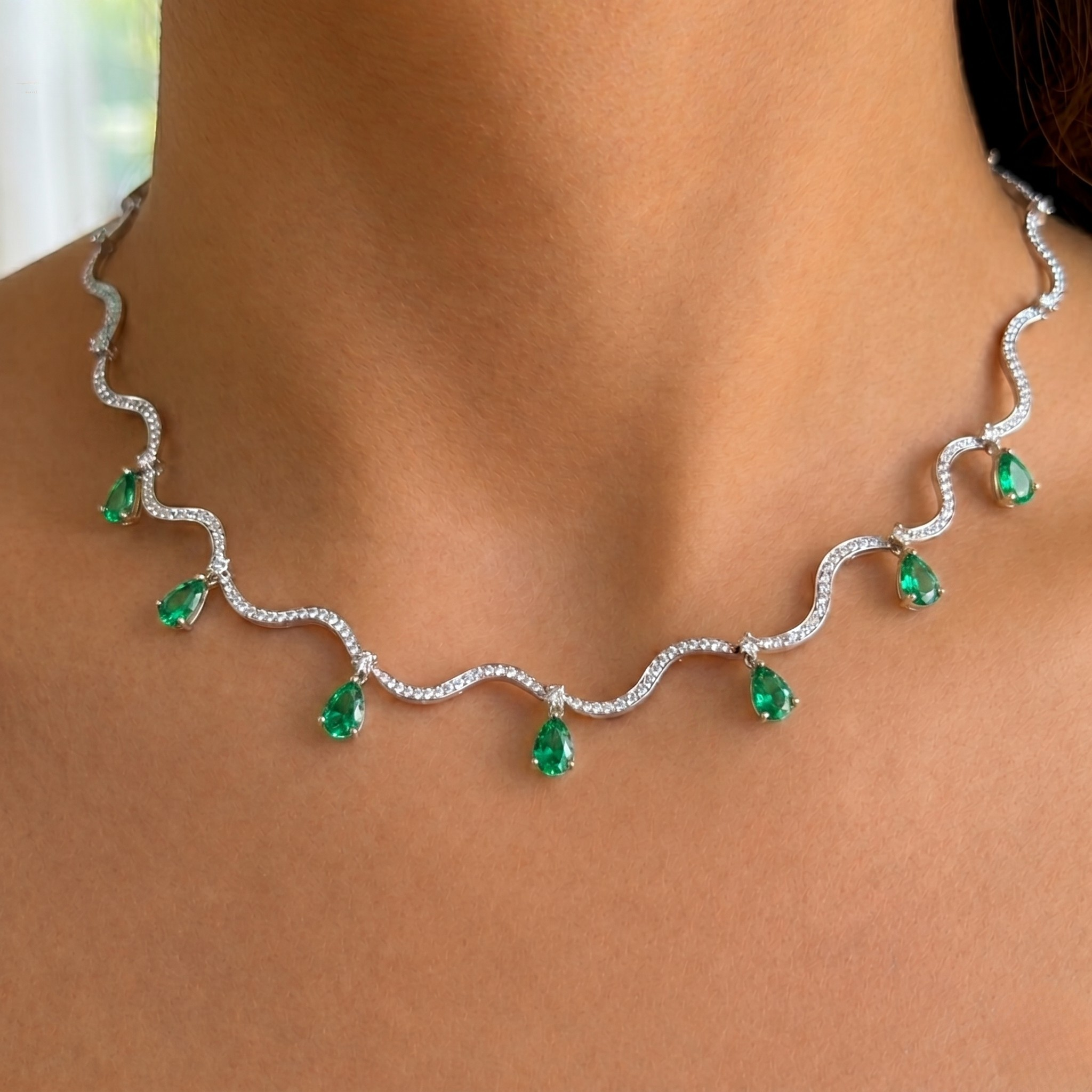 A close-up of a wavy silver-toned necklace with small clear stones and seven green teardrop emerald-cut gemstones, shown worn on a model's neck.