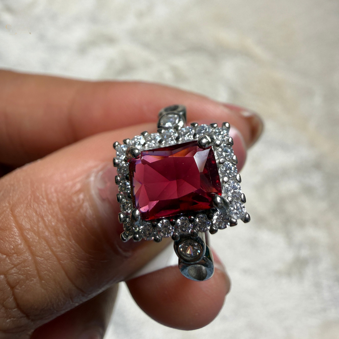 A silver-toned ring featuring a large rectangular red gemstone surrounded by small clear stones in a halo setting, shown being held between fingers against a light textured background.