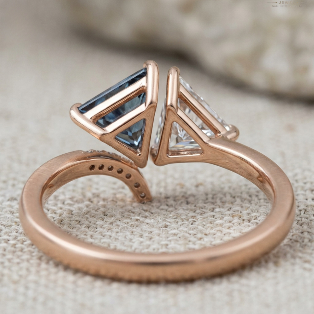 A close-up of a rose gold open-shank ring with a triangular bezel setting featuring a rectangular light blue gemstone and a clear stone, resting on a textured beige fabric background.