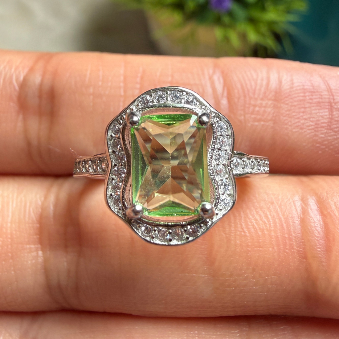A silver-toned halo ring with a large iridescent green and gold cushion-cut center stone, shown worn on a person's finger.