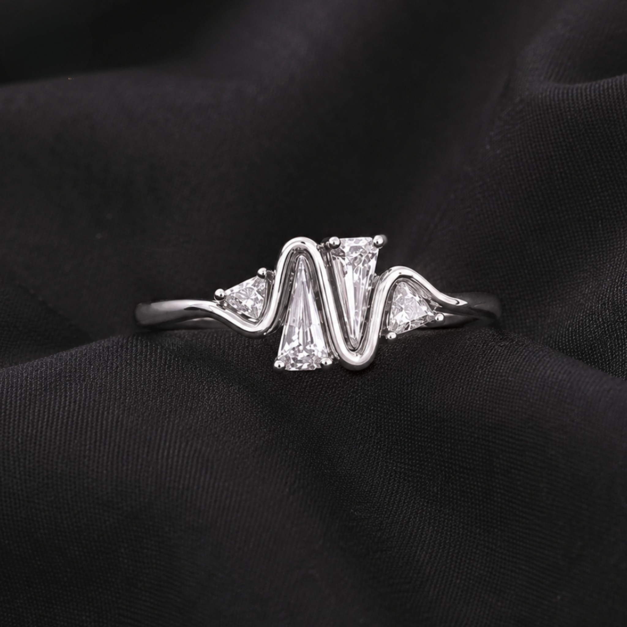 A silver ring with a wavy, zigzag design featuring a large rectangular clear center stone and smaller triangular side stones, displayed on a textured black fabric background.