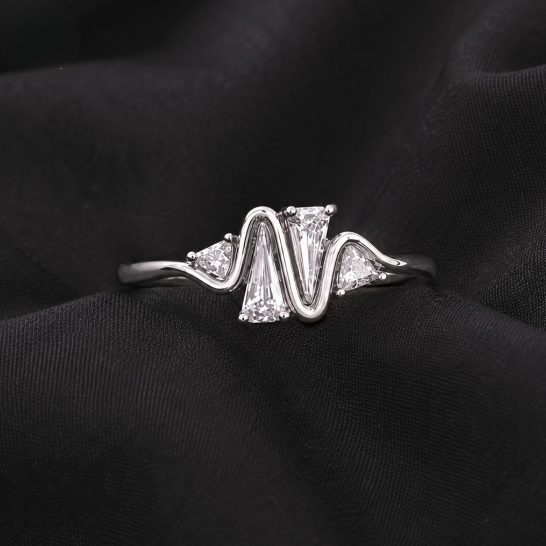 A silver ring with a wavy, zigzag design featuring a large rectangular clear center stone and smaller triangular side stones, displayed on a textured black fabric background.