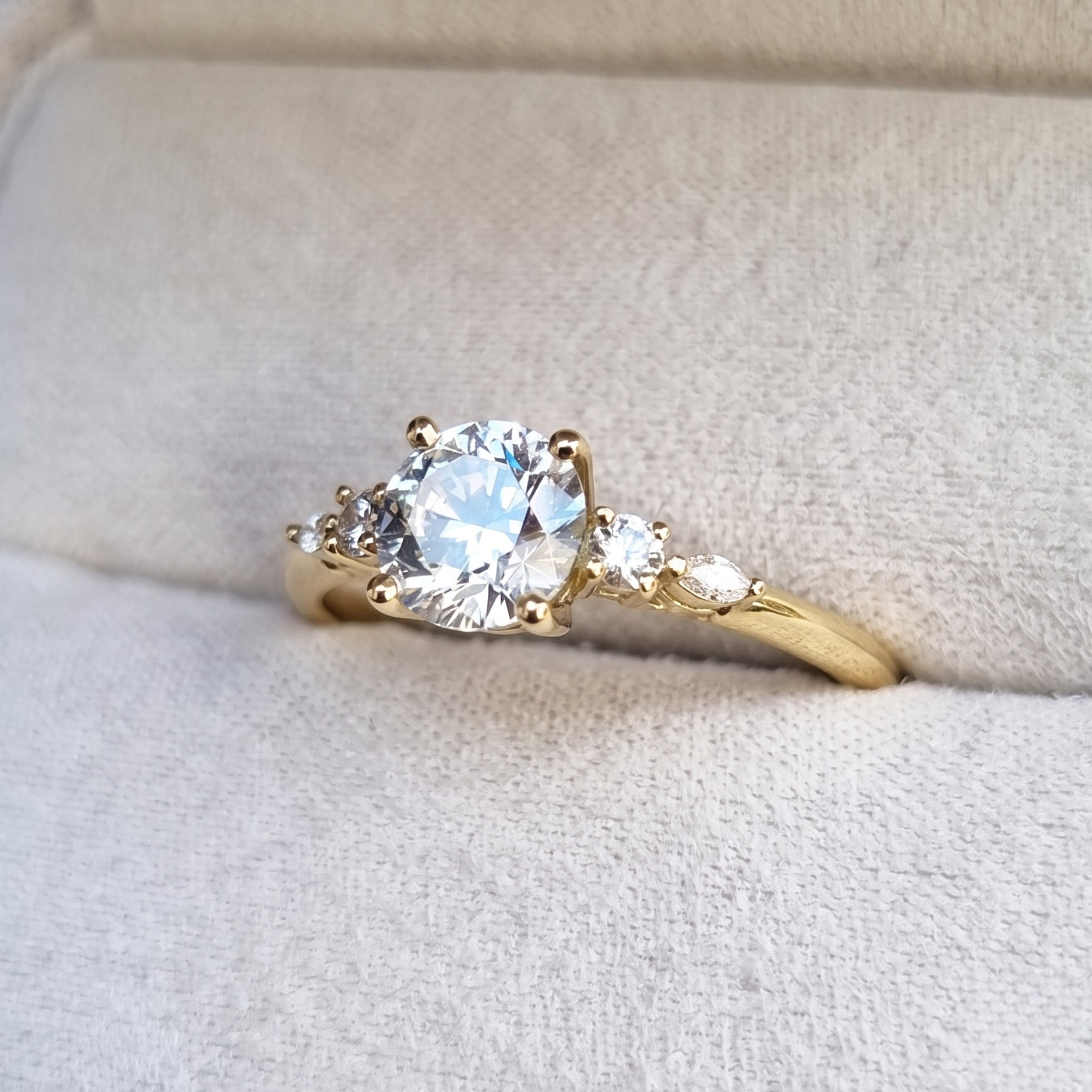 A yellow gold engagement ring with a large round brilliant-cut center stone and two smaller marquise-cut side stones, displayed on a textured cream-colored ring box.