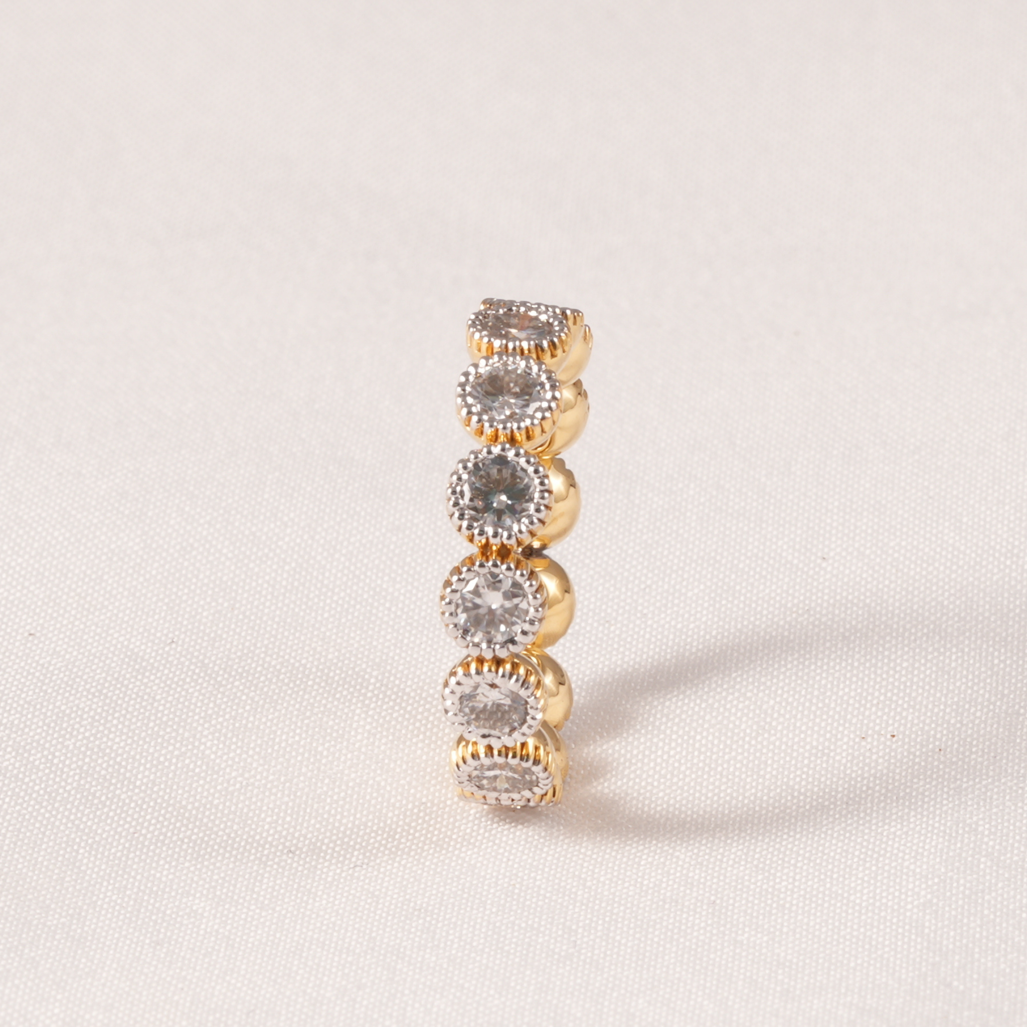 A gold ring featuring five stacked round bezel-set clear gemstones, photographed on a light beige textured fabric background.