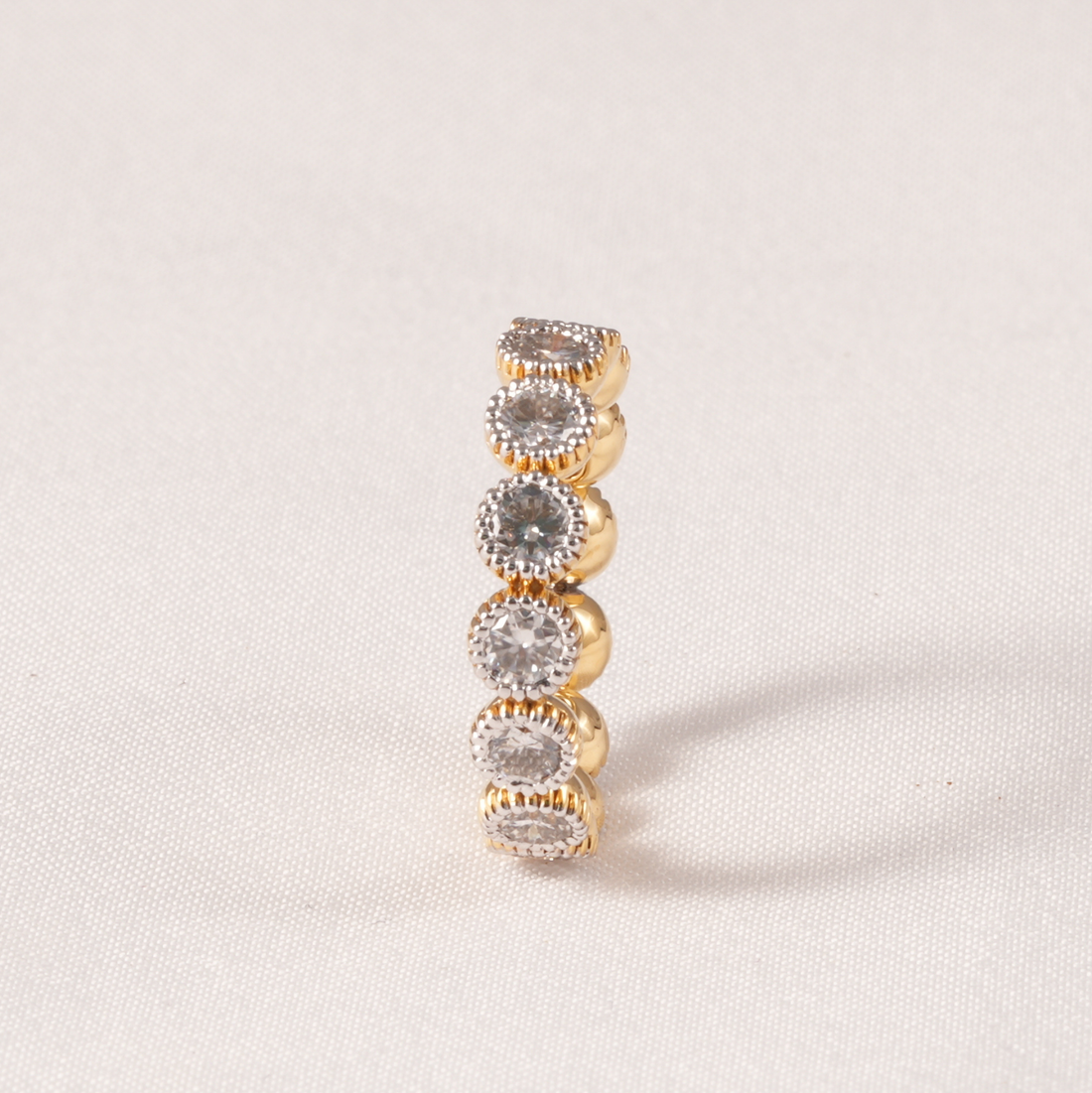 A gold ring featuring five stacked round bezel-set clear gemstones, photographed on a light beige textured fabric background.