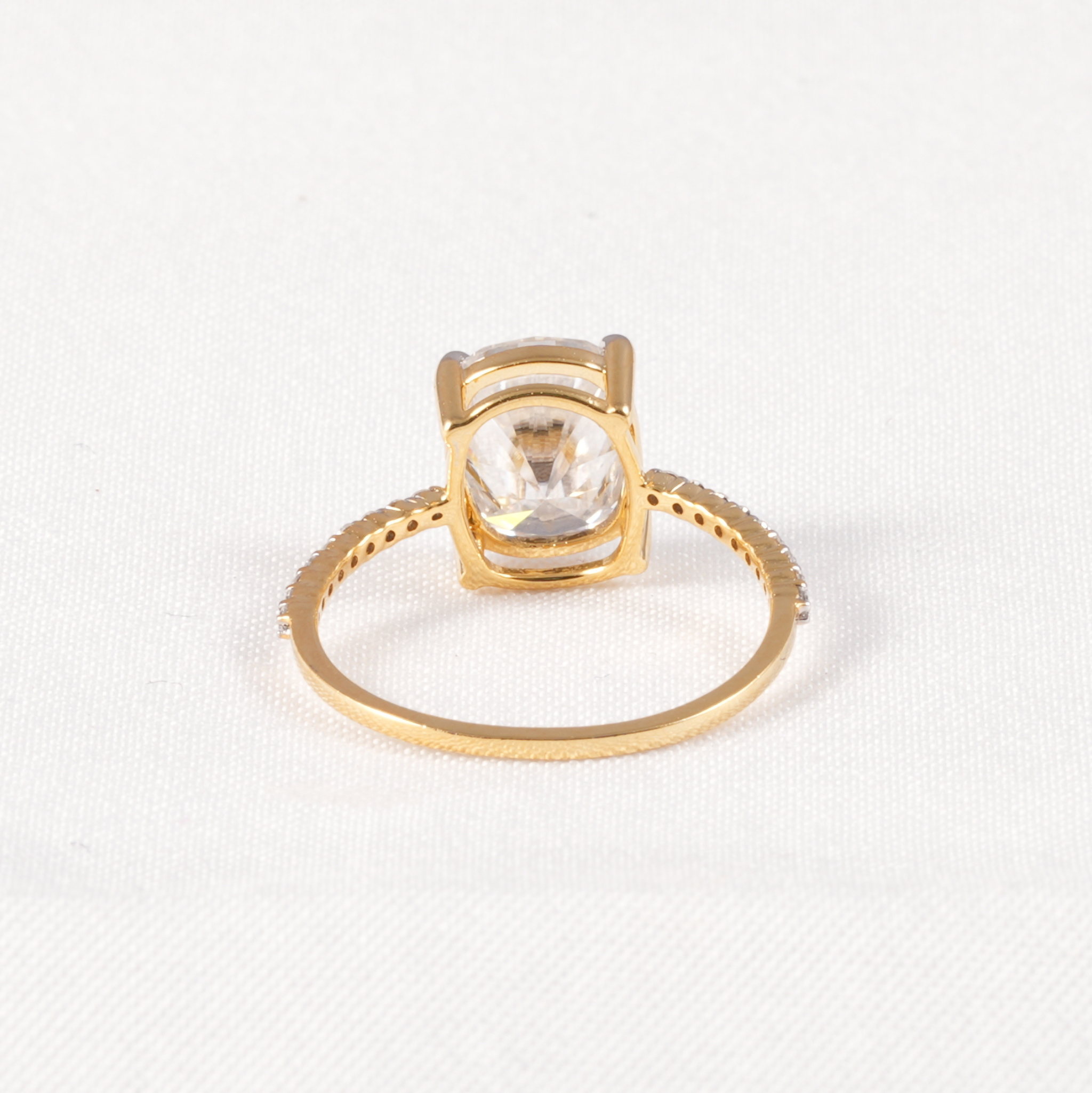 A gold ring with a square bezel setting and a clear round-cut gemstone, shown on a textured white fabric background.