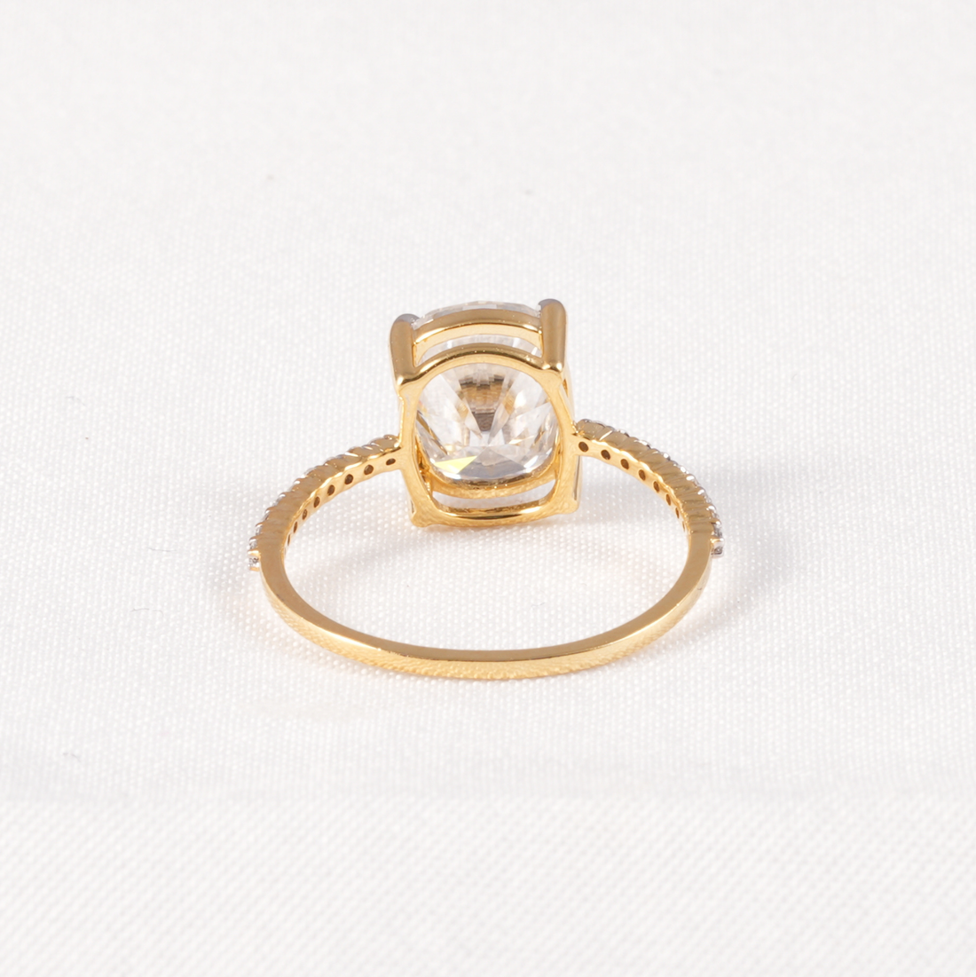 A gold ring with a square bezel setting and a clear round-cut gemstone, shown on a textured white fabric background.