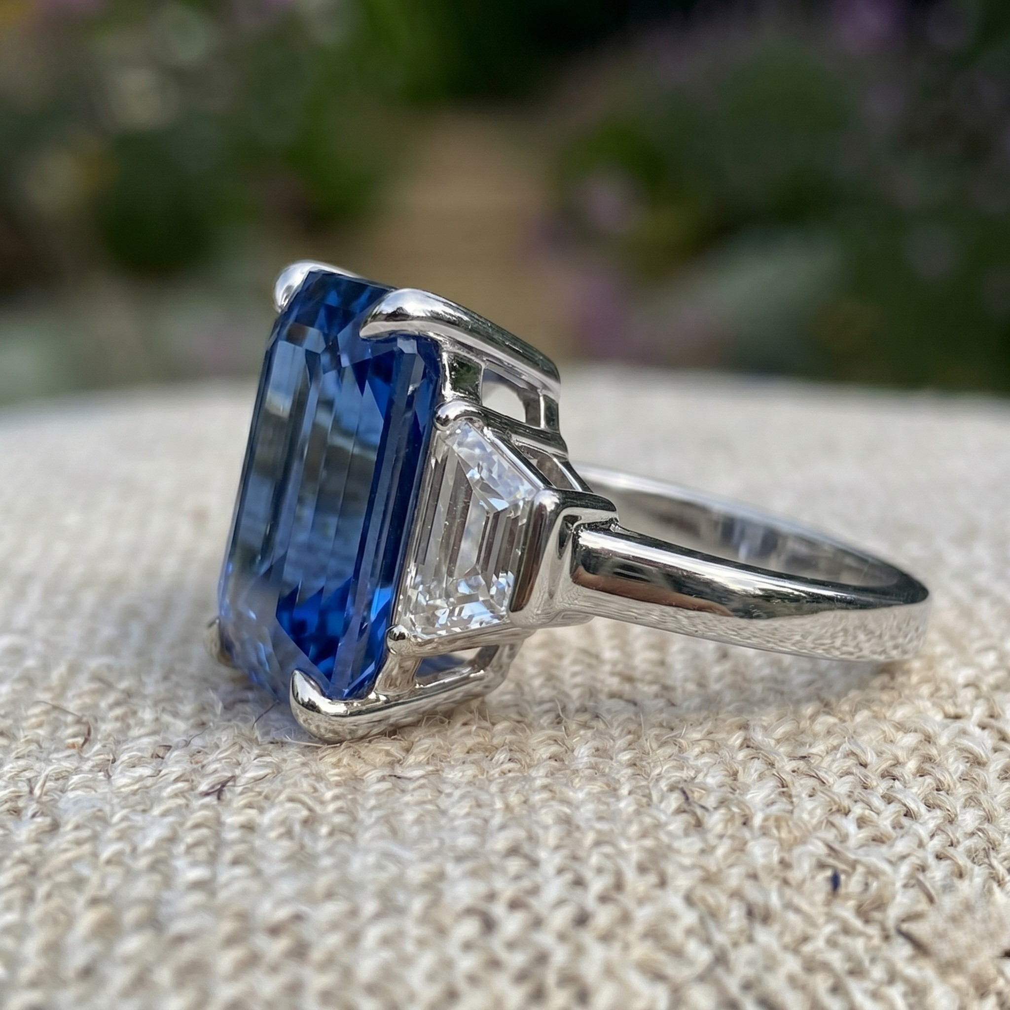A silver ring featuring a large rectangular blue sapphire gemstone and a smaller baguette-cut clear stone, resting on a textured beige fabric with a blurred garden background.