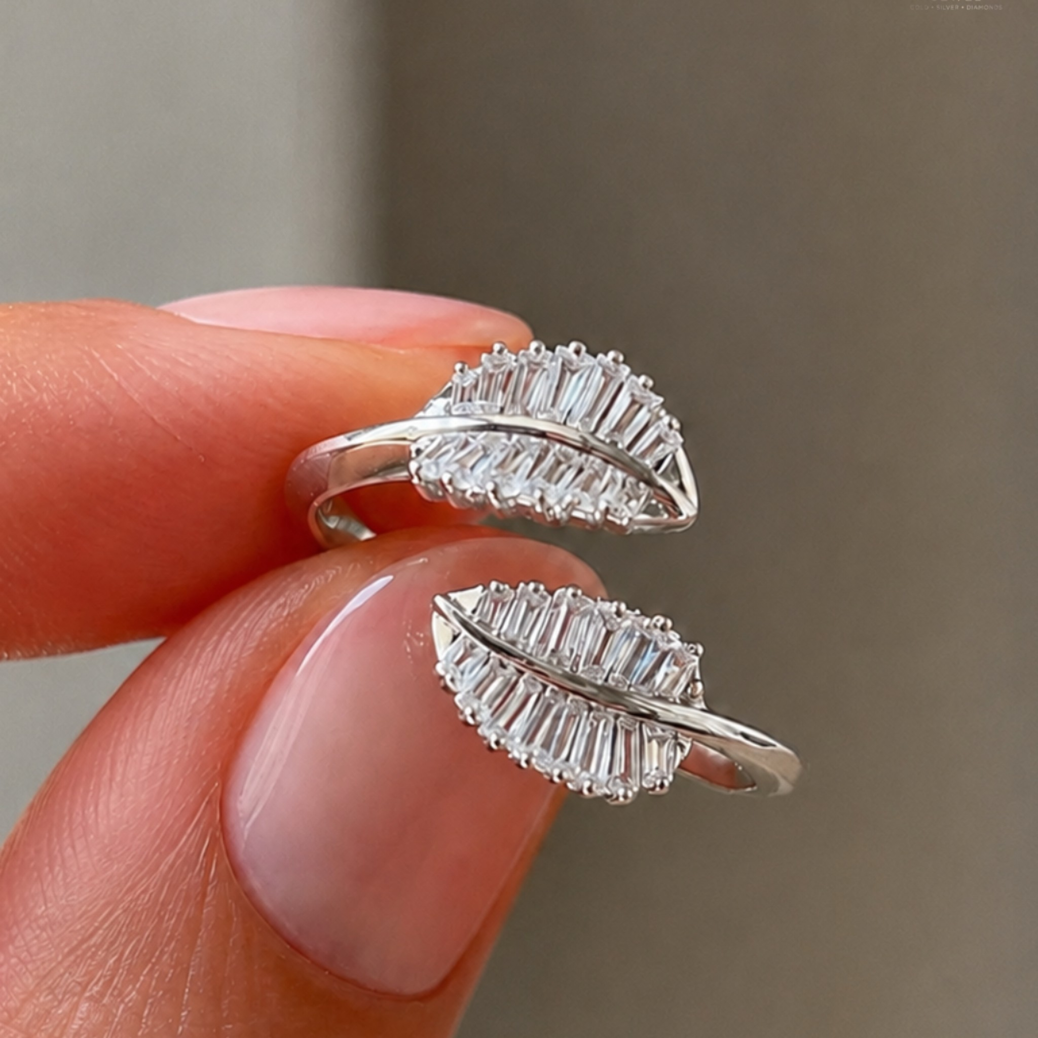 A close-up of a person's fingers holding a pair of silver leaf-shaped rings set with clear baguette gemstones against a soft gray background.