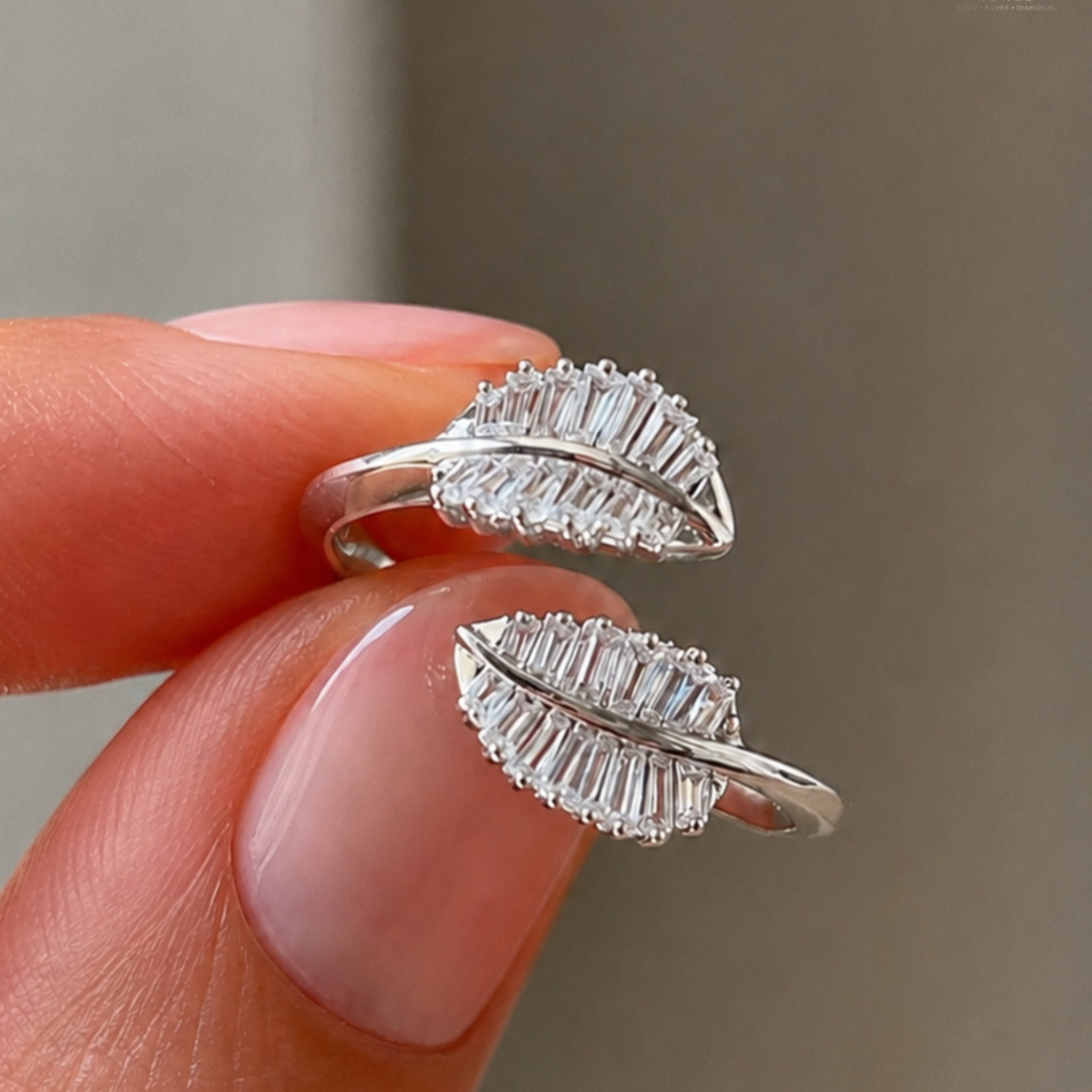 A close-up of a person's fingers holding a pair of silver leaf-shaped rings set with clear baguette gemstones against a soft gray background.