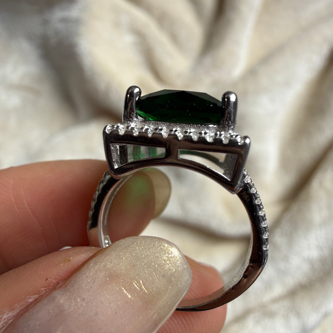 A close-up of a hand holding a silver ring with a large dark green oval center stone and a halo of small clear stones, set against a soft beige fabric background.