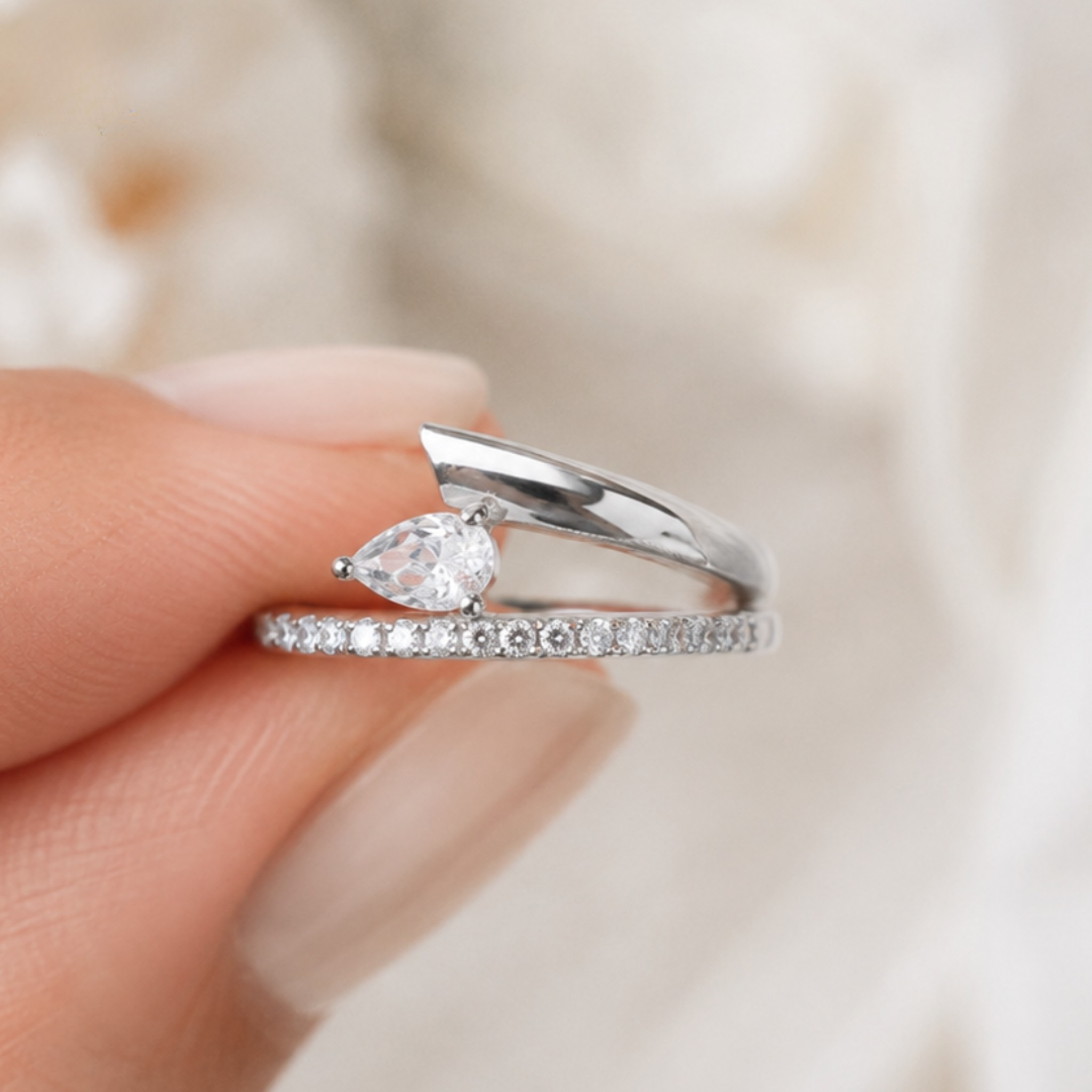 A silver ring with a pear-cut clear gemstone and a pavé band, held between fingers against a soft, out-of-focus cream background.