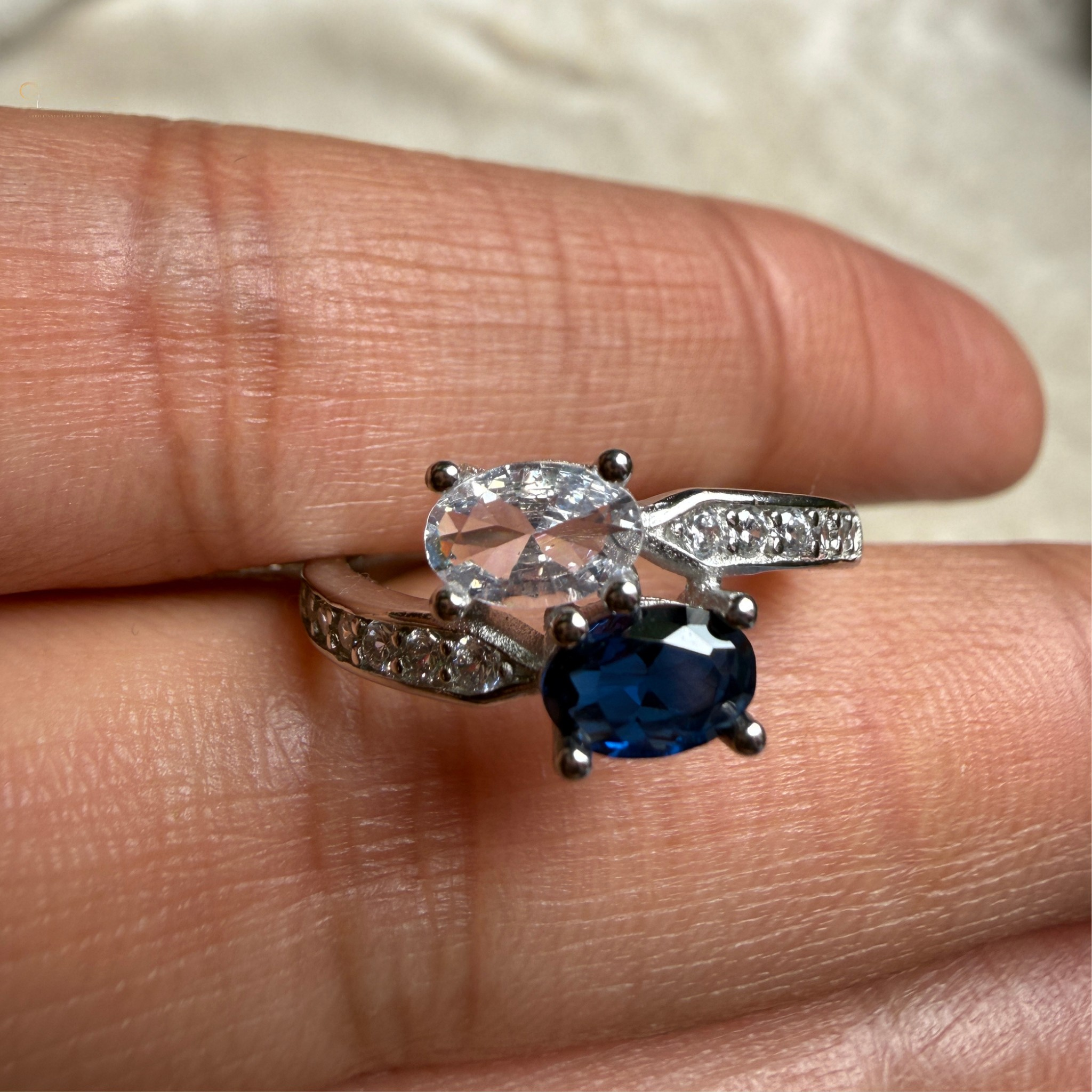 A silver-tone ring with an oval-cut clear gemstone and a larger oval-cut deep blue gemstone, shown worn on a person's finger.