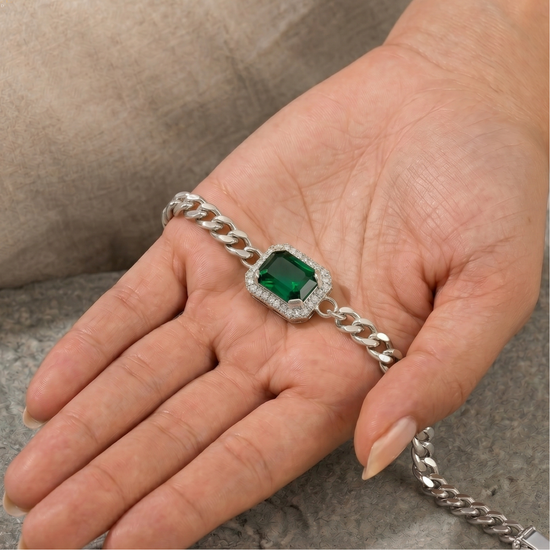 A silver chain bracelet with a rectangular green emerald-cut center stone surrounded by a halo of small clear stones, displayed on a person's hand.