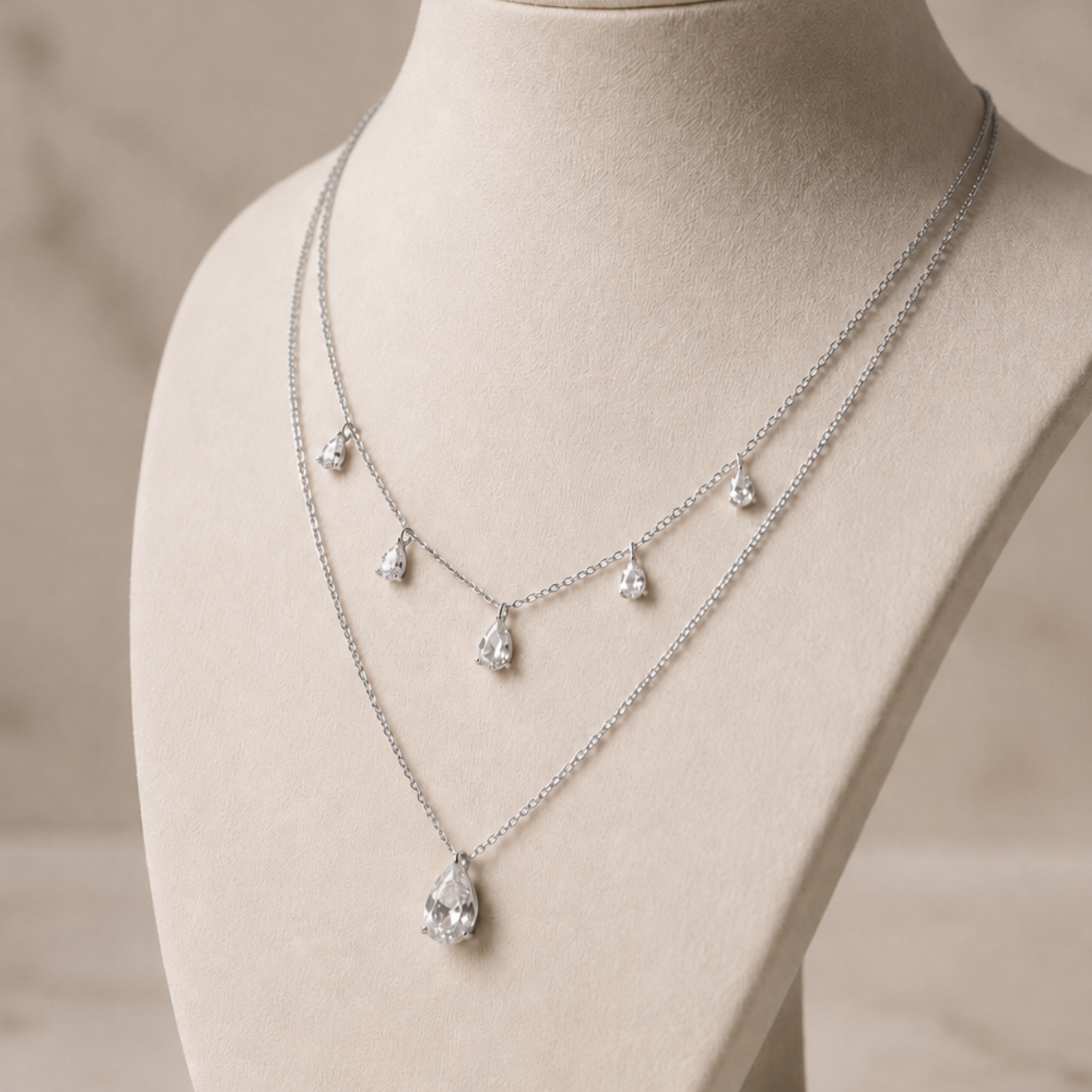 A double-layered silver necklace with clear teardrop-shaped gemstones, displayed on a beige jewelry bust against a soft neutral background.