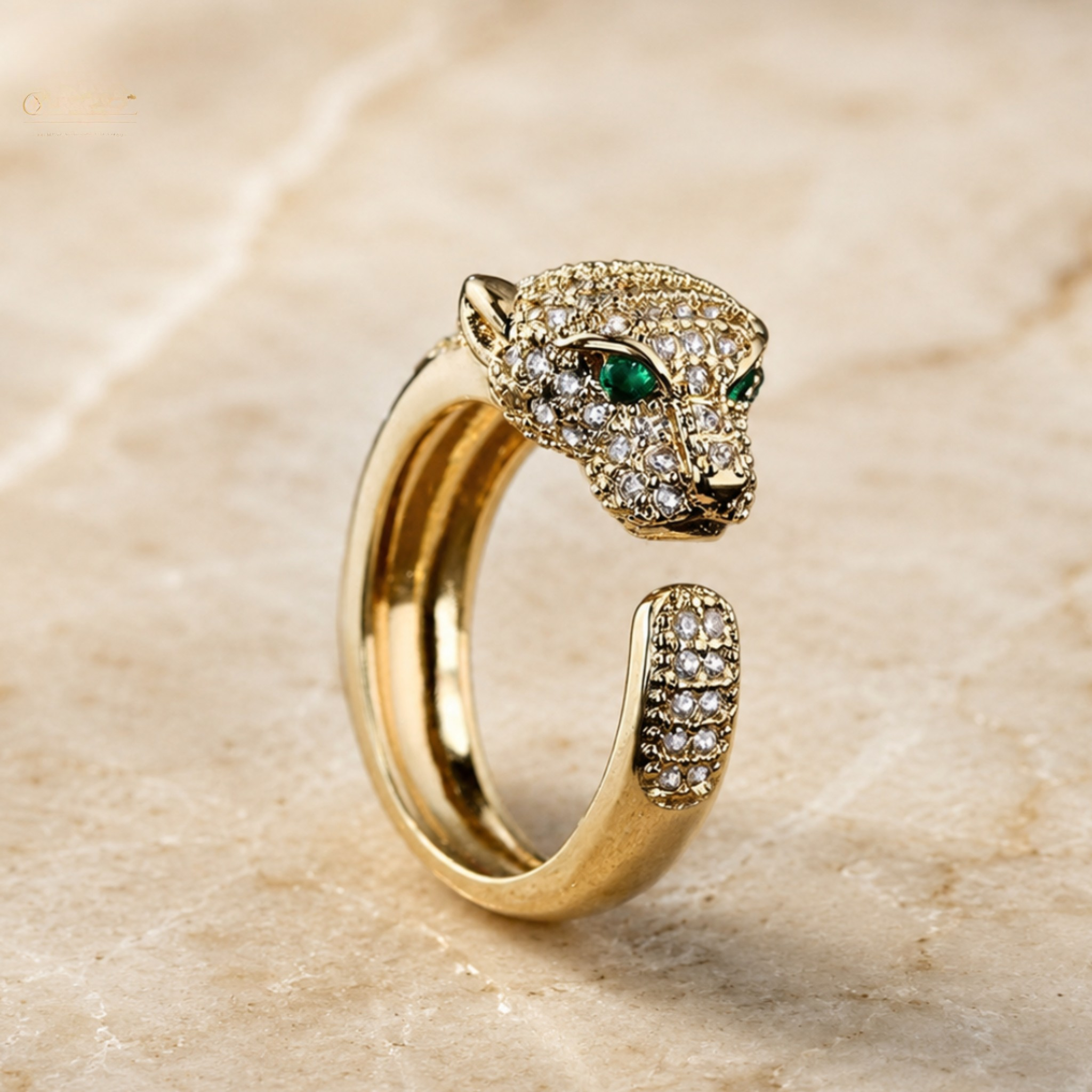 A gold-toned open ring designed like a panther head, featuring pavé-set clear stones and green gemstone eyes, displayed on a beige marble surface.
