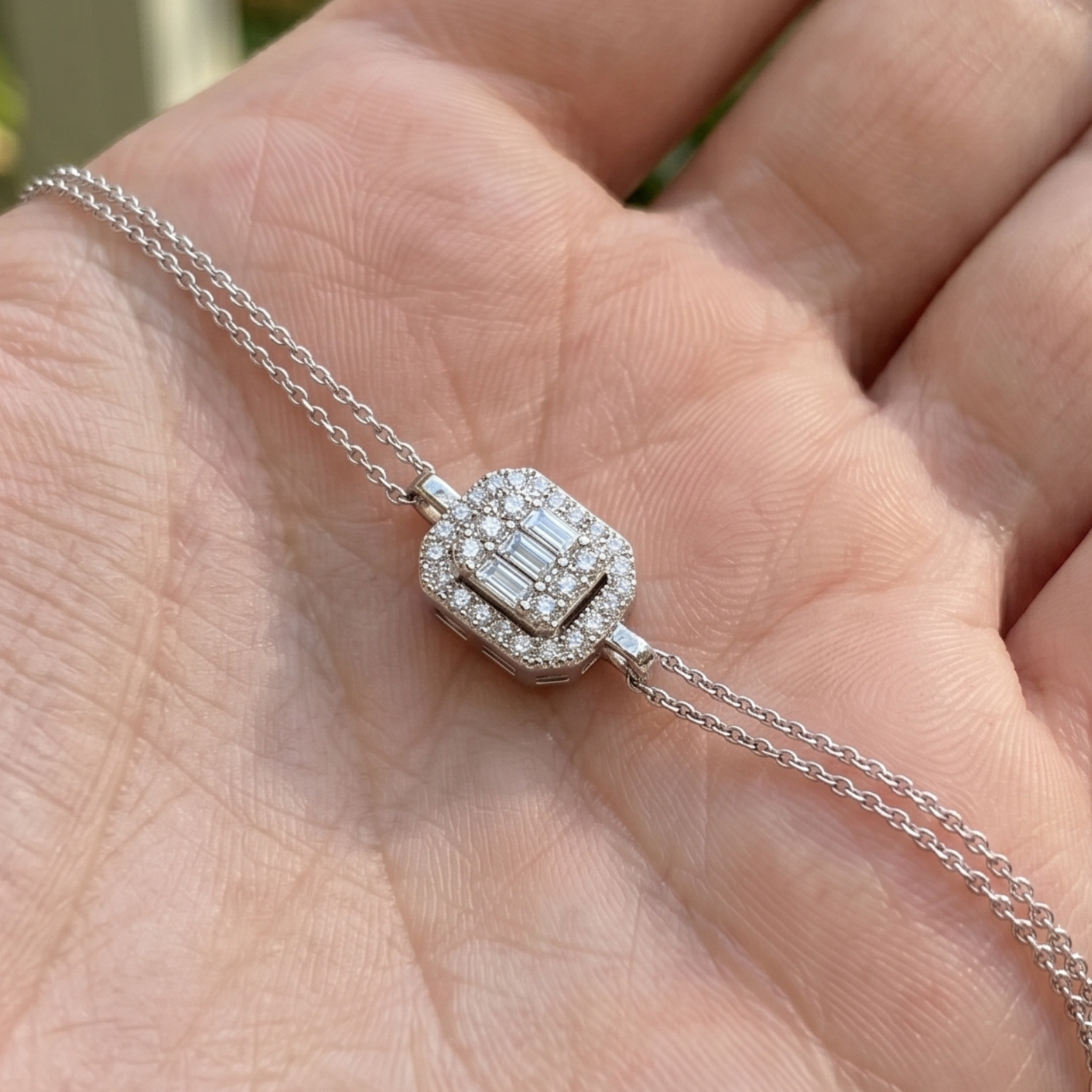A close-up of a silver-toned diamond necklace with a rectangular baguette-cut center stone and a halo of small diamonds, resting on an open hand.