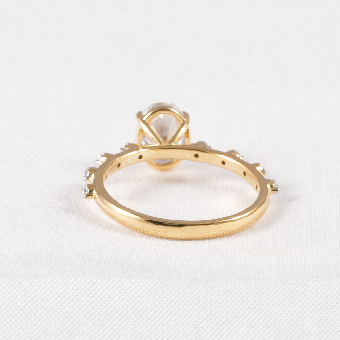 A yellow gold ring with a large clear gemstone in a prong setting and smaller clear stones along the band, photographed on a white textured fabric background.
