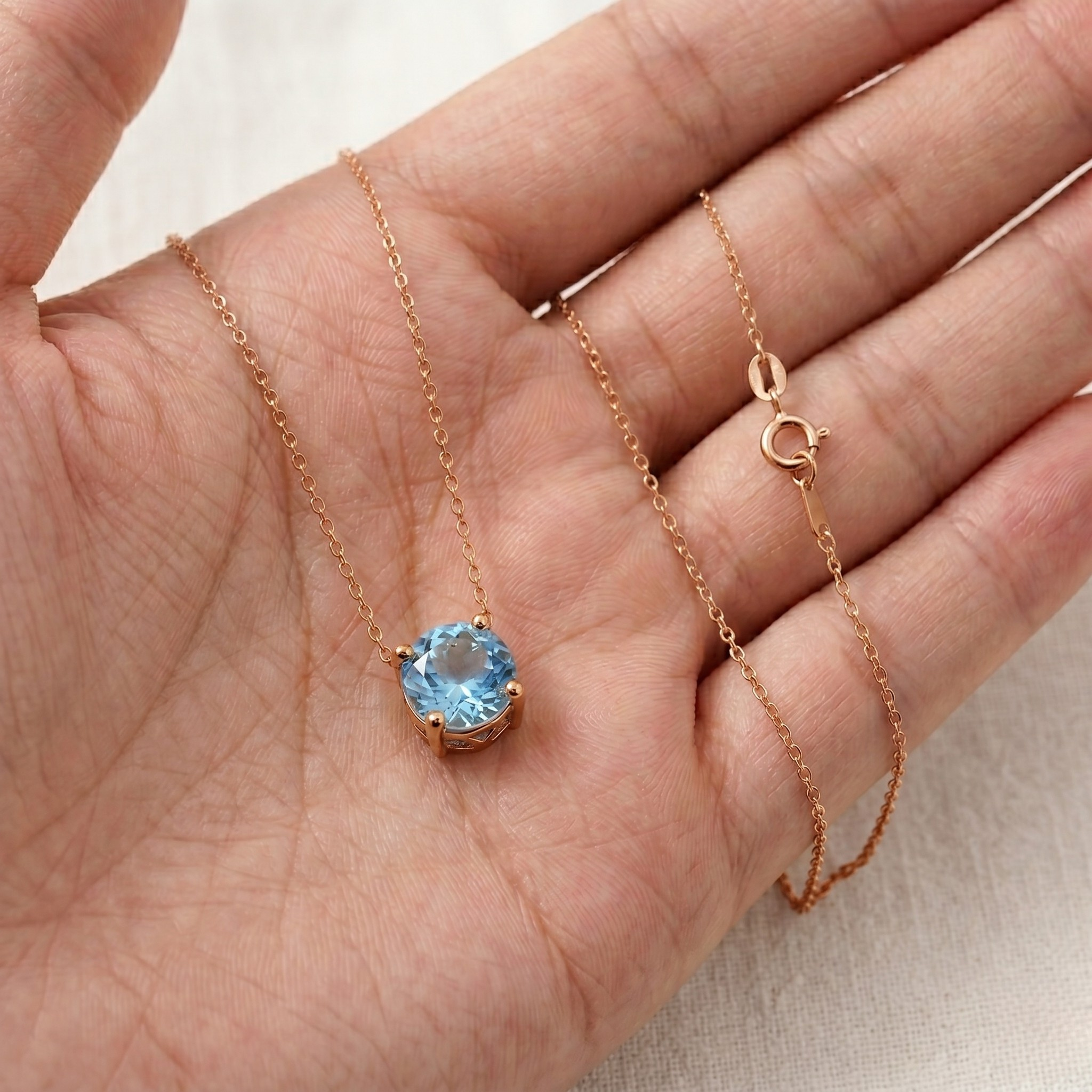 A delicate rose gold necklace with a round-cut light blue gemstone pendant, displayed on an open hand against a soft beige fabric background.