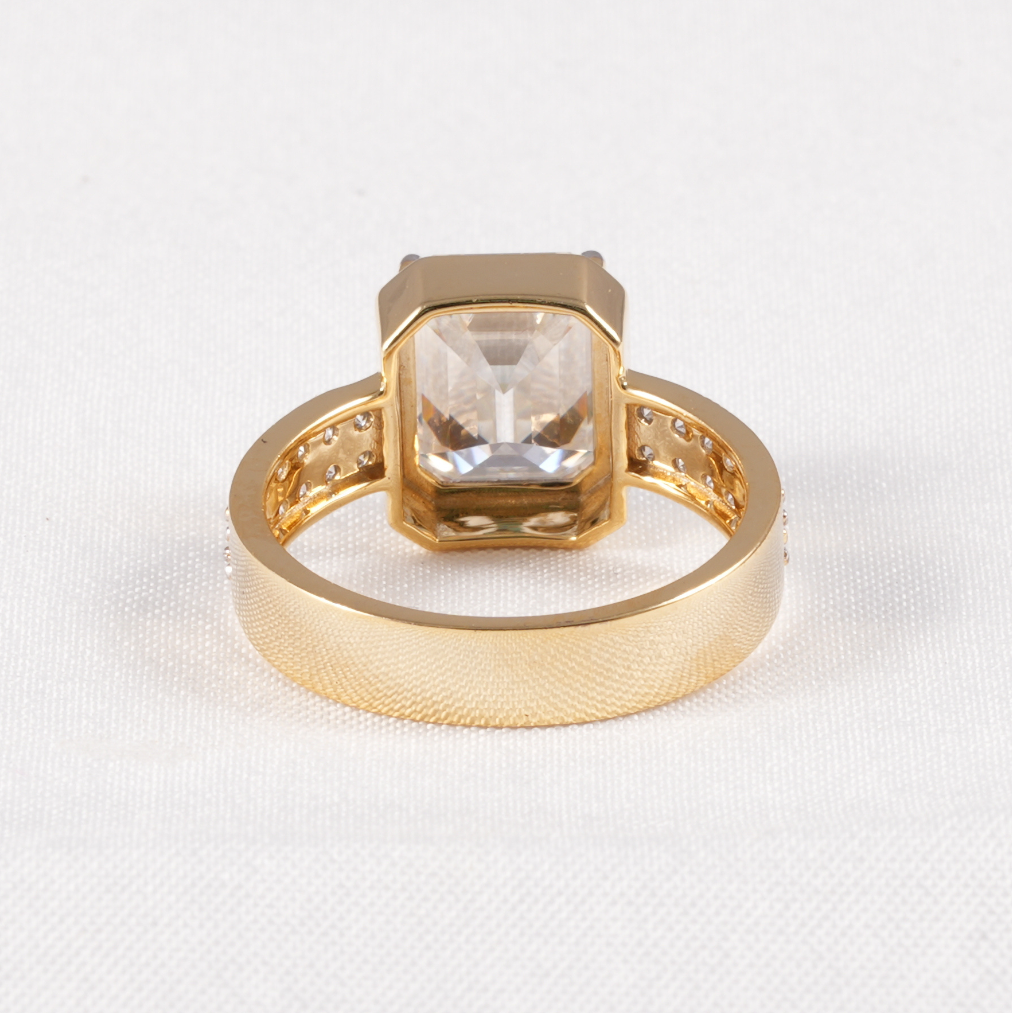 A gold ring featuring a large, square-cut clear gemstone in a bezel setting, with small accent stones along the band, photographed on a textured white fabric background.