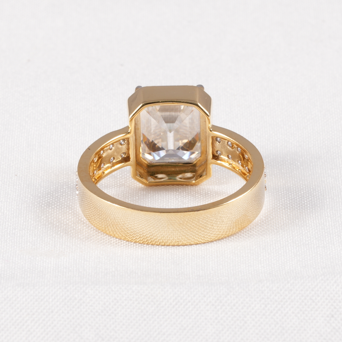 A gold ring featuring a large, square-cut clear gemstone in a bezel setting, with small accent stones along the band, photographed on a textured white fabric background.
