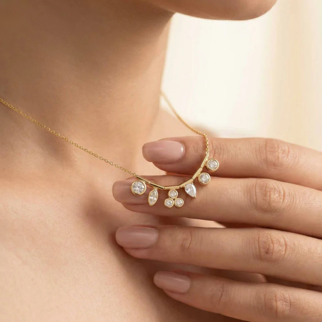 A close-up lifestyle shot of a delicate gold crescent necklace with clear teardrop and round gemstones, worn on a model's neck and held gently by her hand.