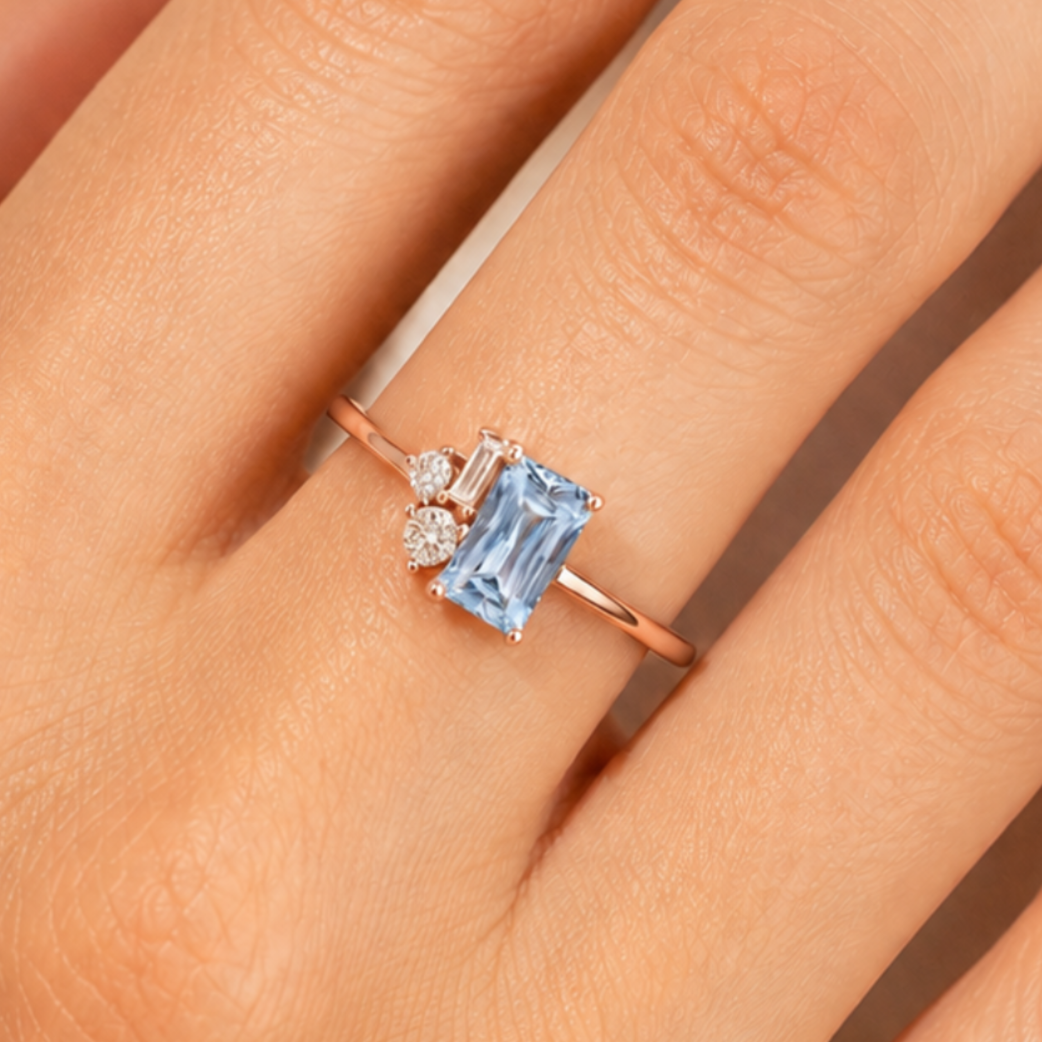 A close-up of a rose gold ring with a rectangular light blue gemstone and a small clear accent stone, worn on a finger.