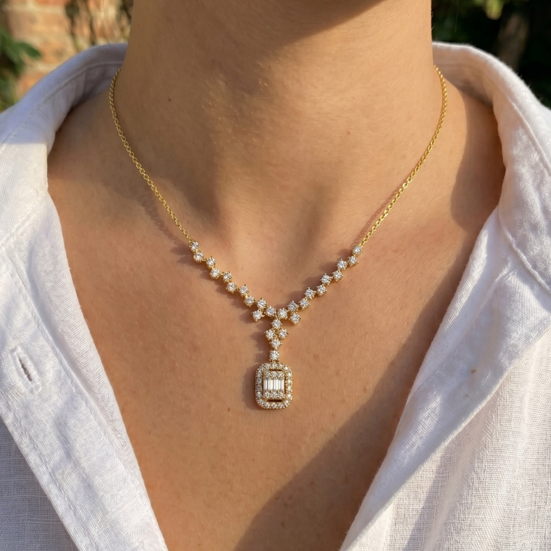 A close-up of a person wearing a delicate gold necklace with a rectangular pendant set with clear stones, styled with a white collared shirt.
