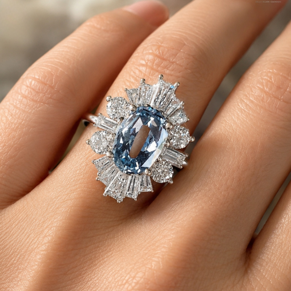 A close-up of a hand wearing a silver ring featuring a large oval light blue gemstone surrounded by a halo of baguette and round diamonds.