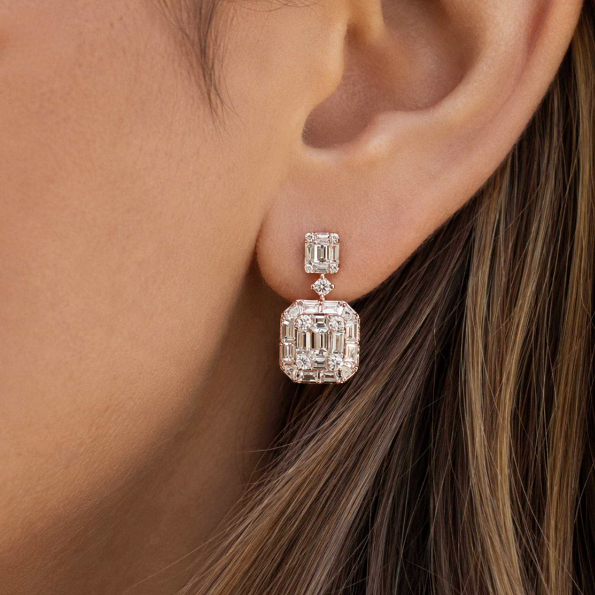 A close-up of a person's ear wearing a silver drop earring with a large emerald-cut clear gemstone and a smaller matching stone above it.