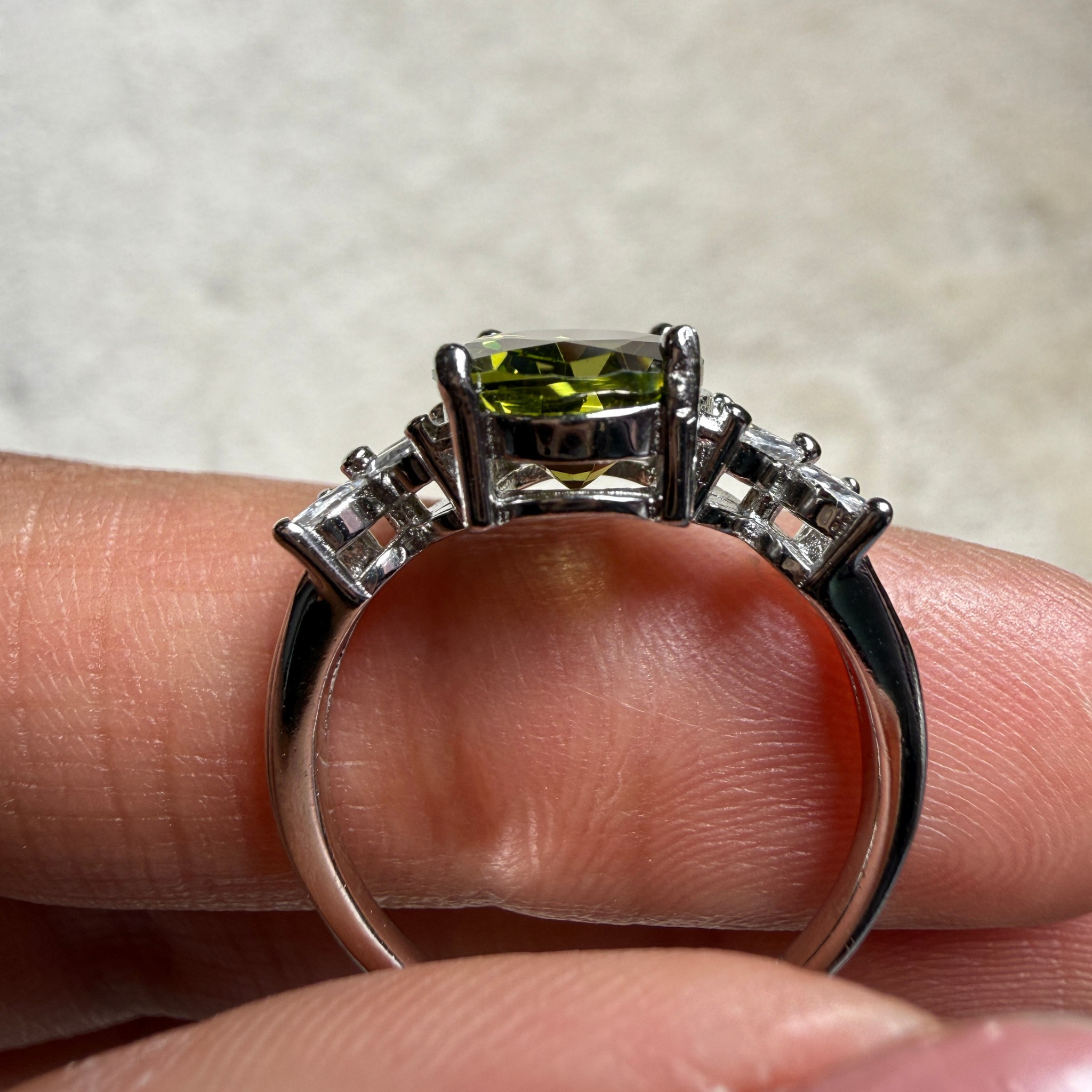 A silver-toned ring with a rectangular green gemstone and smaller side stones, shown resting on a person's fingers against a light neutral background.