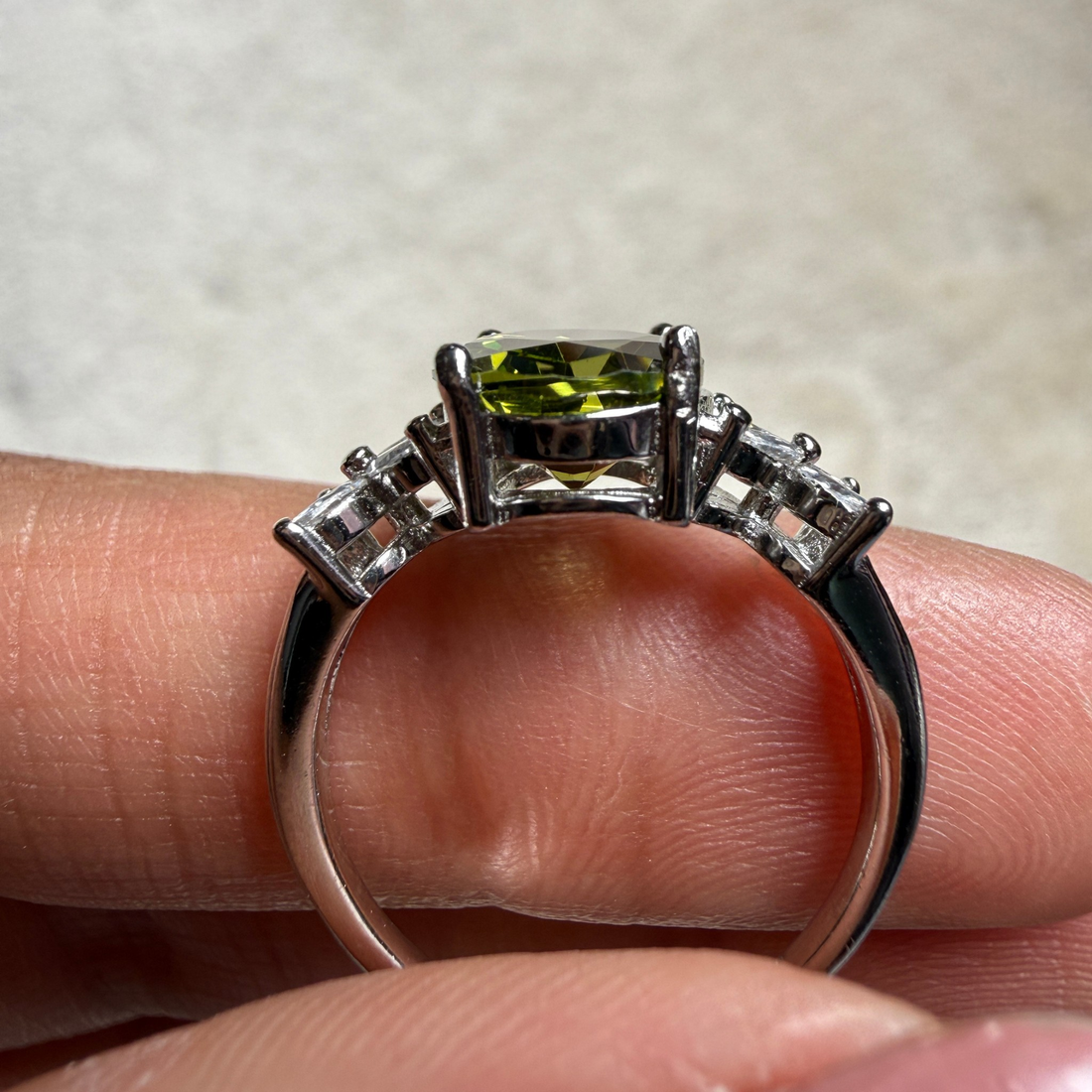 A silver-toned ring with a rectangular green gemstone and smaller side stones, shown resting on a person's fingers against a light neutral background.