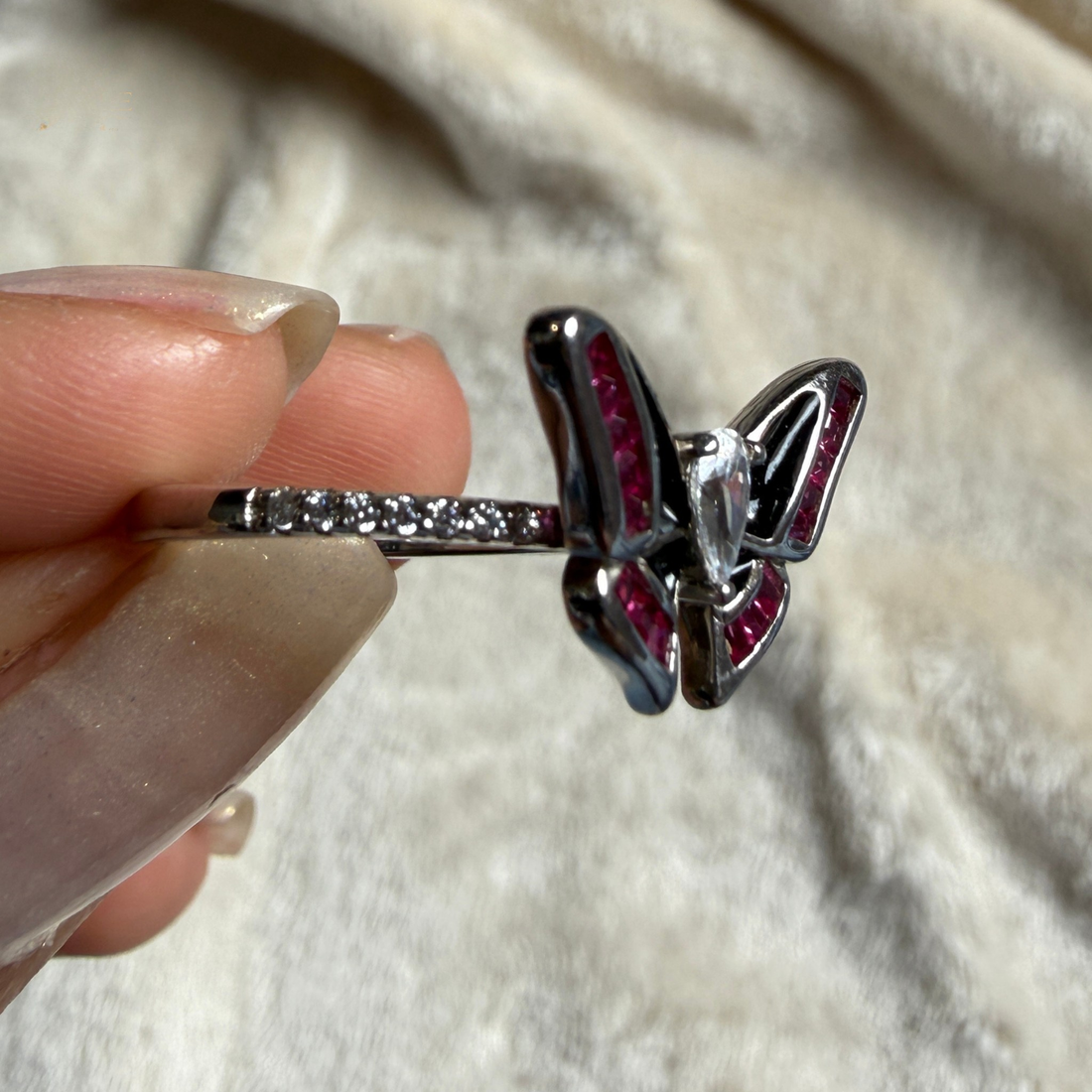 A silver butterfly ring with dark red gemstone accents, held between fingers against a soft beige fabric background.