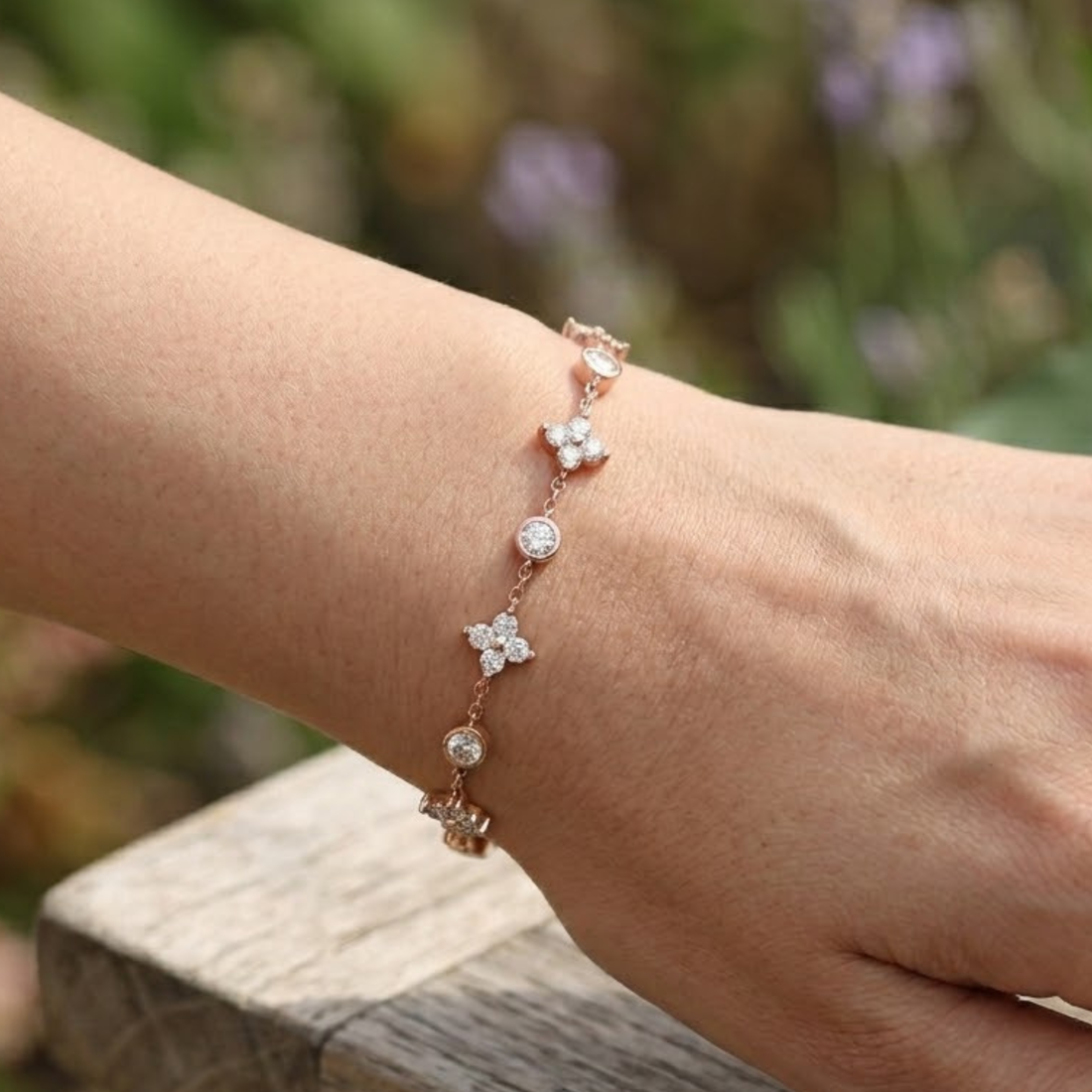 A delicate rose gold chain bracelet with small floral and circular crystal charms, shown worn on a person's wrist against a blurred outdoor background.