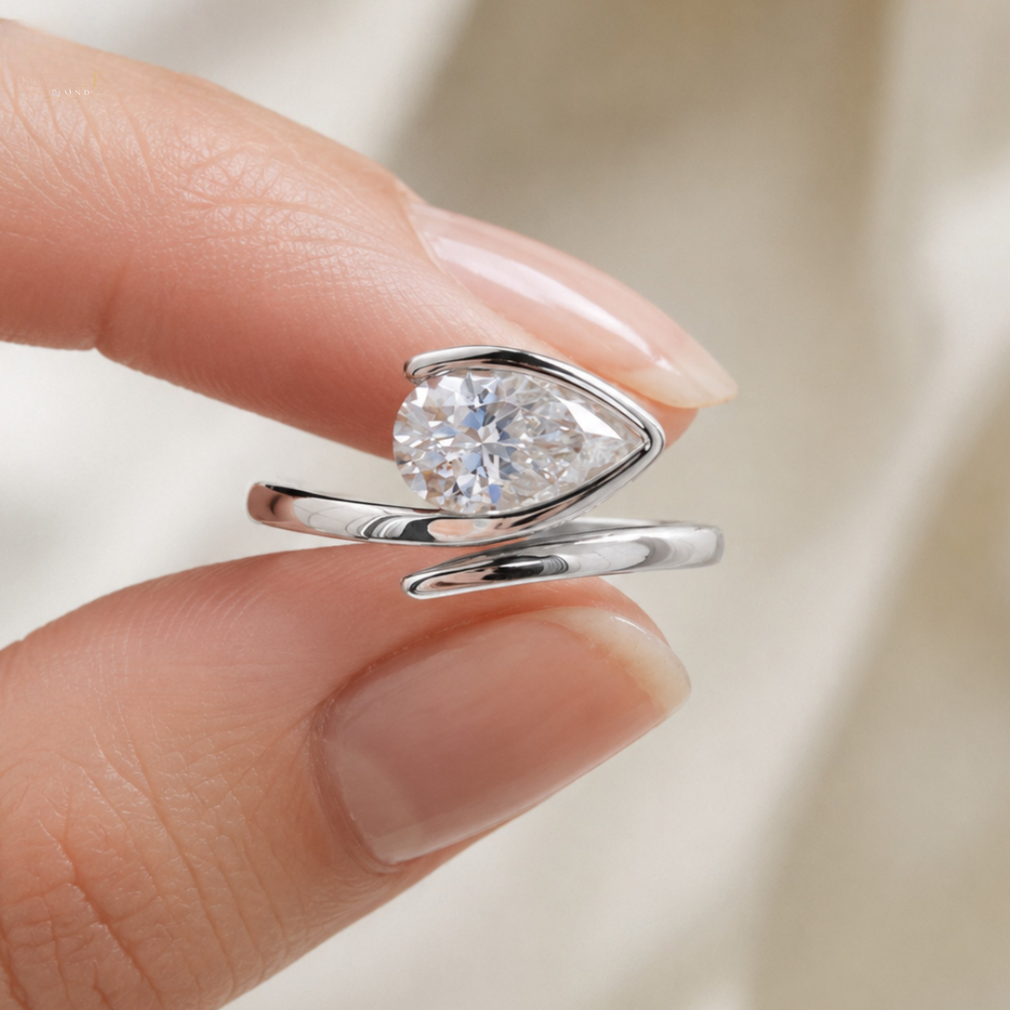 A silver-tone ring with a pear-cut clear gemstone held between two fingers against a soft, light beige background.