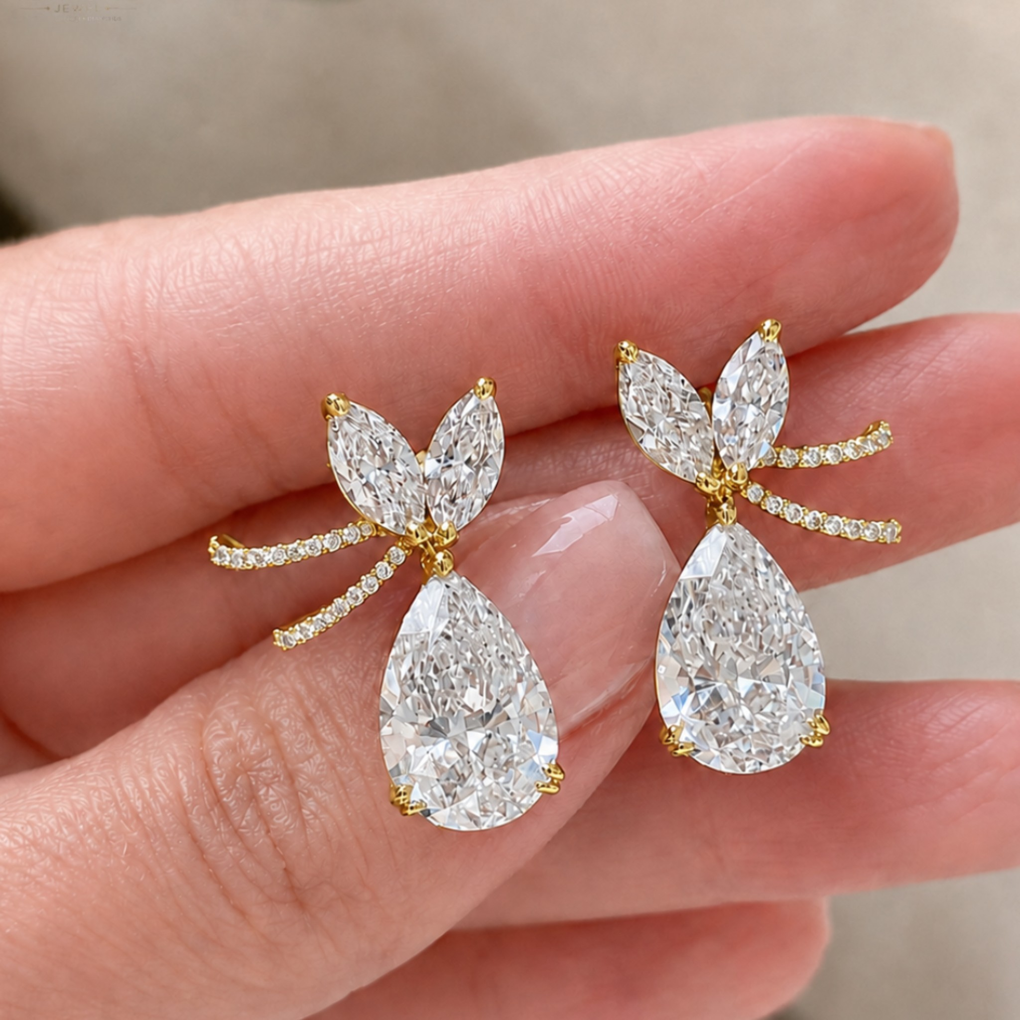 A pair of gold and diamond drop earrings held on a person's fingers. Each earring features a teardrop-shaped clear gemstone with smaller marquise and round-cut clear stones in a yellow gold setting.